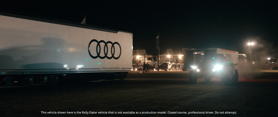 Nachtaufnahme eines Rallye-Fahrzeugs mit Audi-Logo mit Scheinwerfern in einer Campsite bei Dunkelheit.