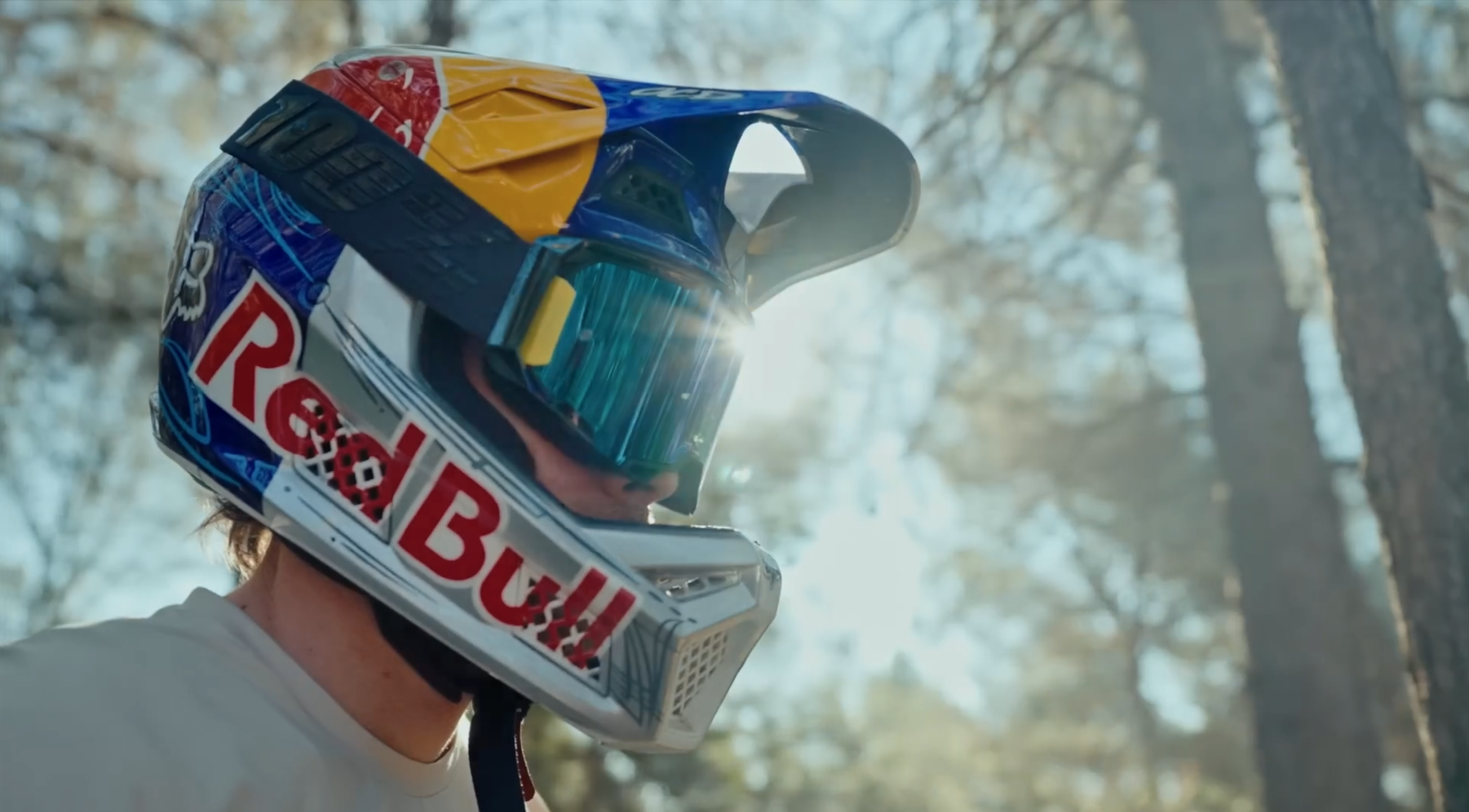 Person trägt einen motocross-Helm mit Red Bull-Logo, Sonnenlicht und Bäume im Hintergrund.