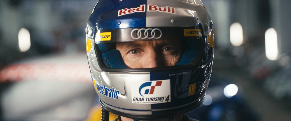 Ein Rennfahrer trägt einen Helm mit Logos von Red Bull, Audi und Gran Turismo 4, blickt durch die Visieröffnung.