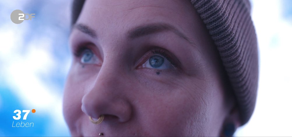 Nahaufnahme eines lächelnden Mädchens mit blauen Augen, Nasenpiercing, beiger Mütze bei Sonnenlicht.