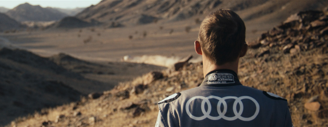 Ein Mann in einem Rennfahrer-Anzug blickt auf eine alpine Landschaft mit Bergen im Hintergrund.