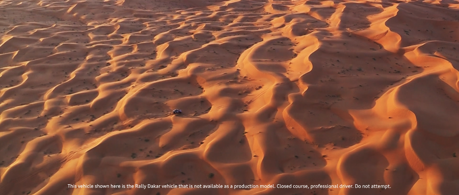 Wüstenlandschaft mit Dünen bei Sonnenuntergang, ein einzelnes Fahrzeug fährt auf Sandwegen.