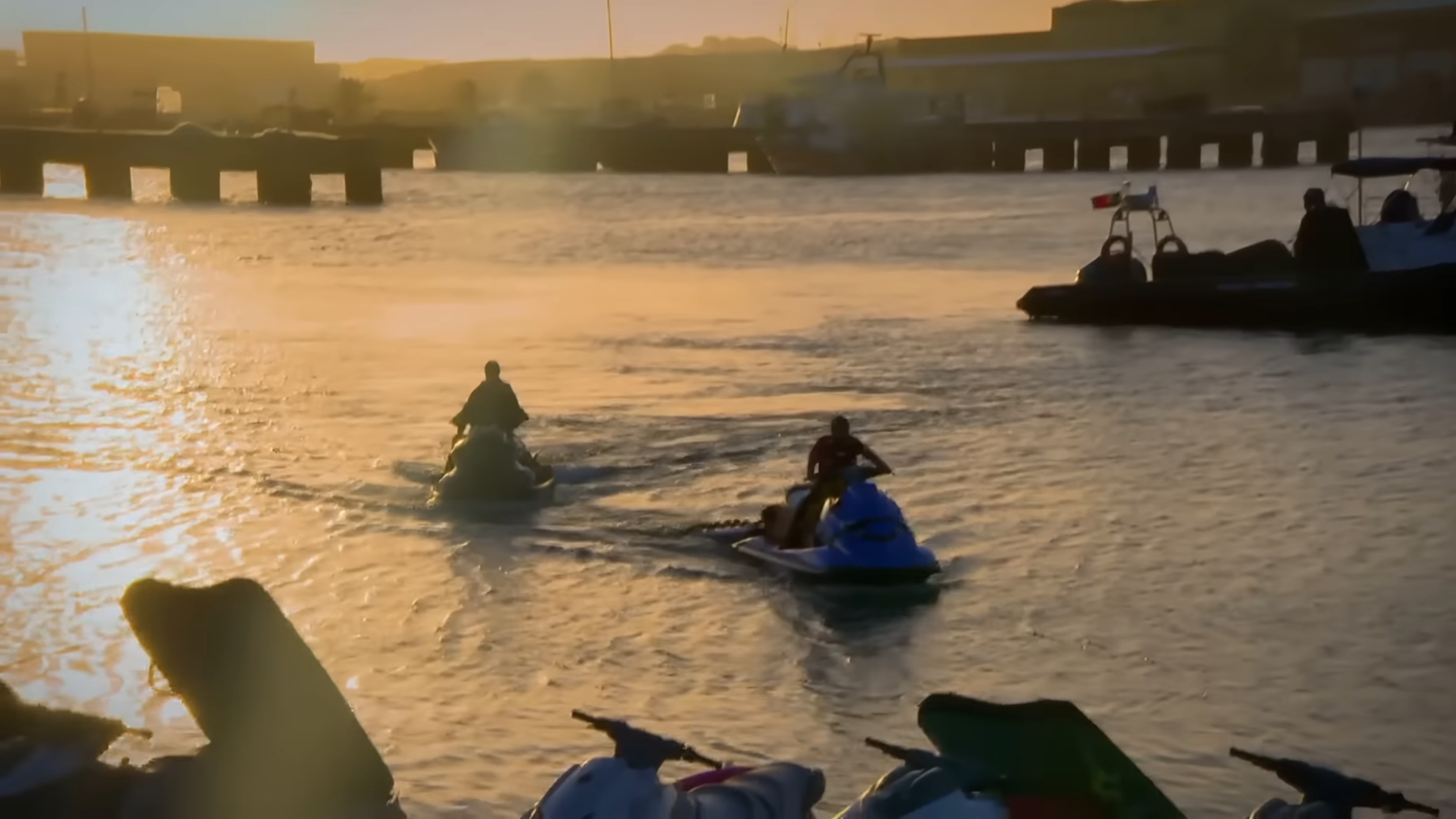 Menschen fahren mit Jet-Skis bei Sonnenuntergang auf einem Wasserweg. Im Hintergrund sind Brücken und Gebäude sichtbar.