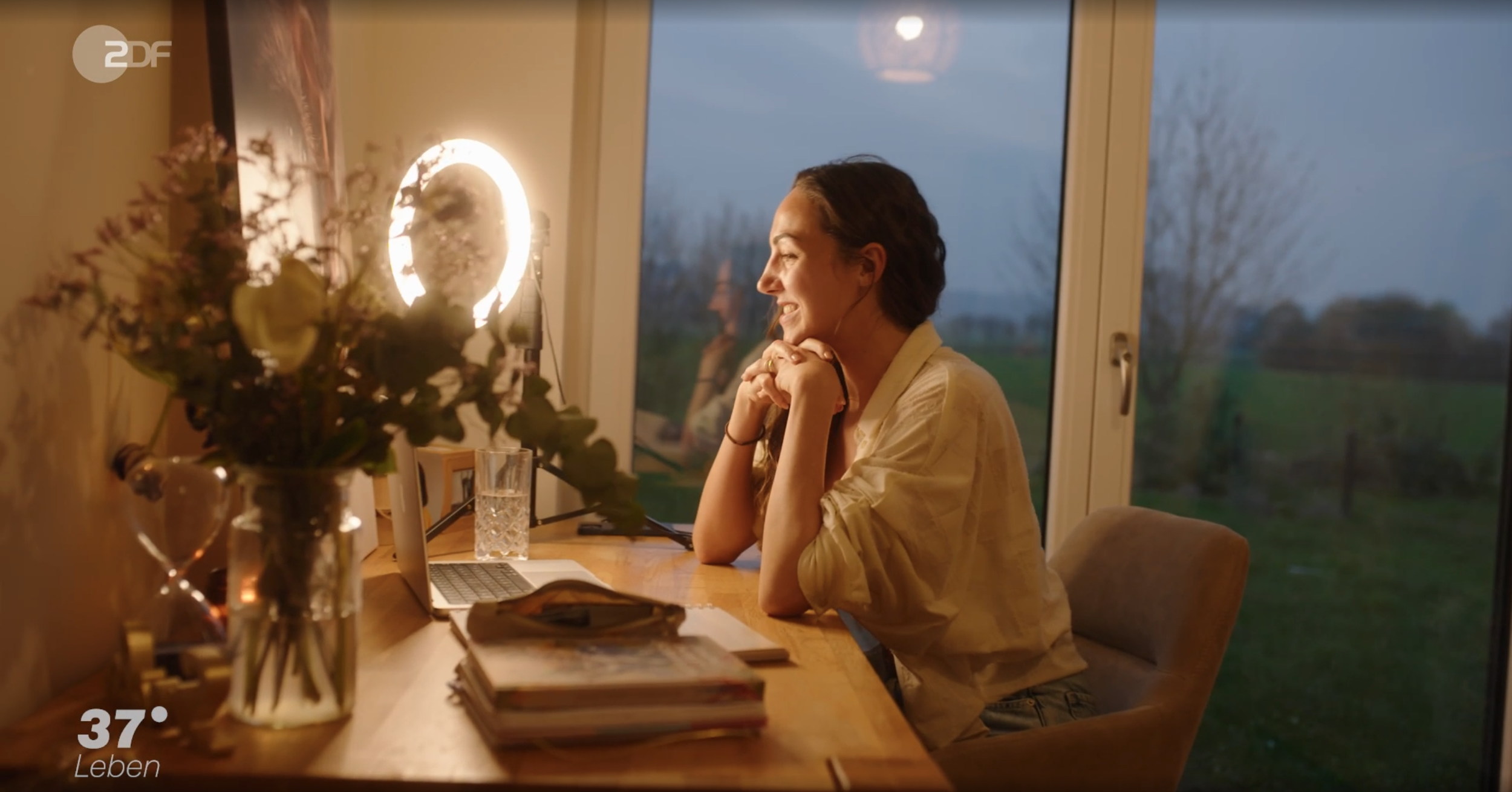 Eine Frau sitzt an einem Tisch vor einem Laptop, im Hauslicht und draußen ist es dunkel. Es ist ein ruhiger Abend. Auf dem Tisch liegen Bücher, und im Hintergrund ist ein Fenster mit Blick nach draußen. Es ist gemütlich und entspannt.