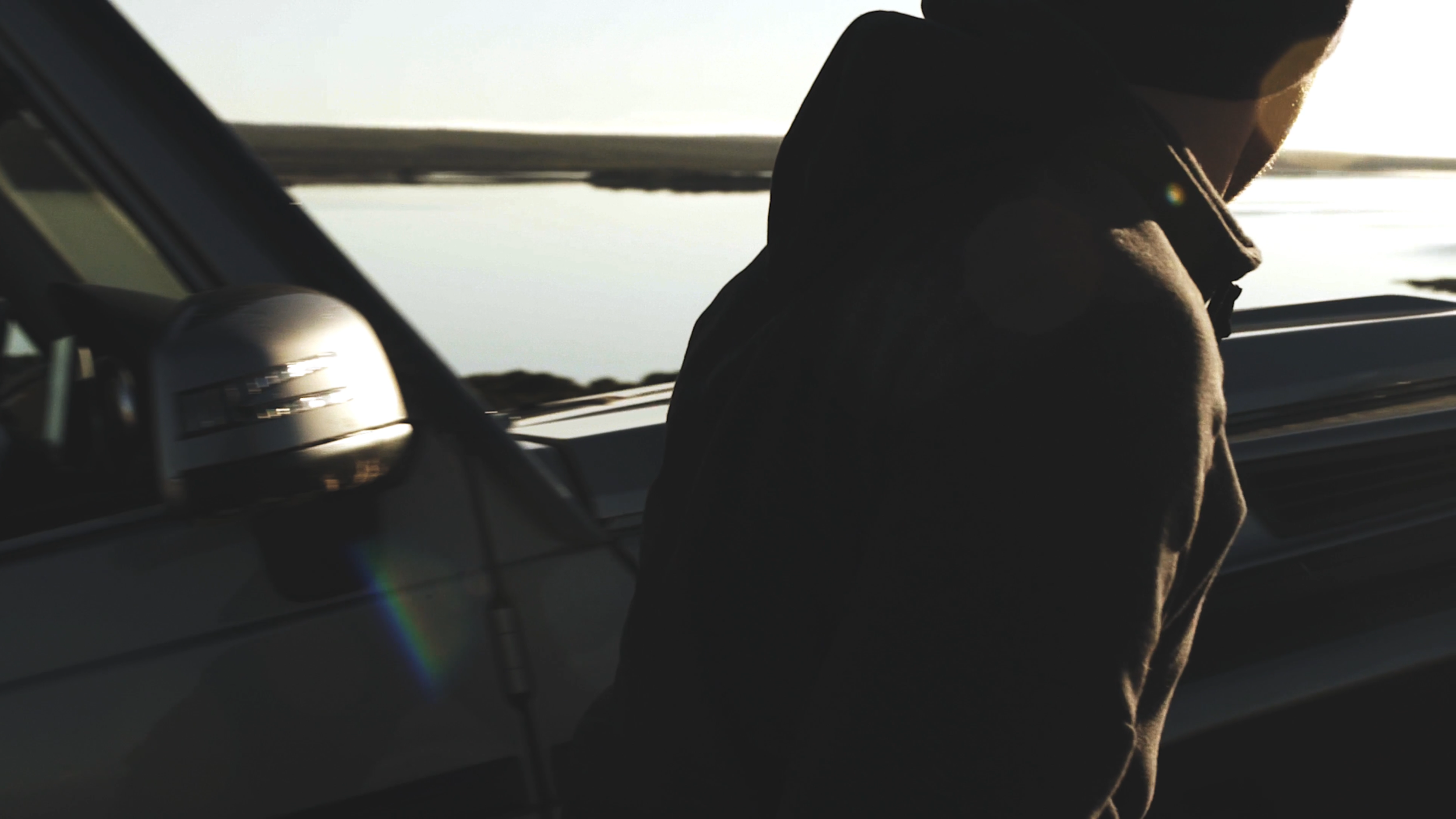 Person sitzt im Auto, Blick nach draußen bei Sonnenuntergang oder Sonnenaufgang, dunkle Silhouette im Inneren des Autos, Meer oder See im Hintergrund.