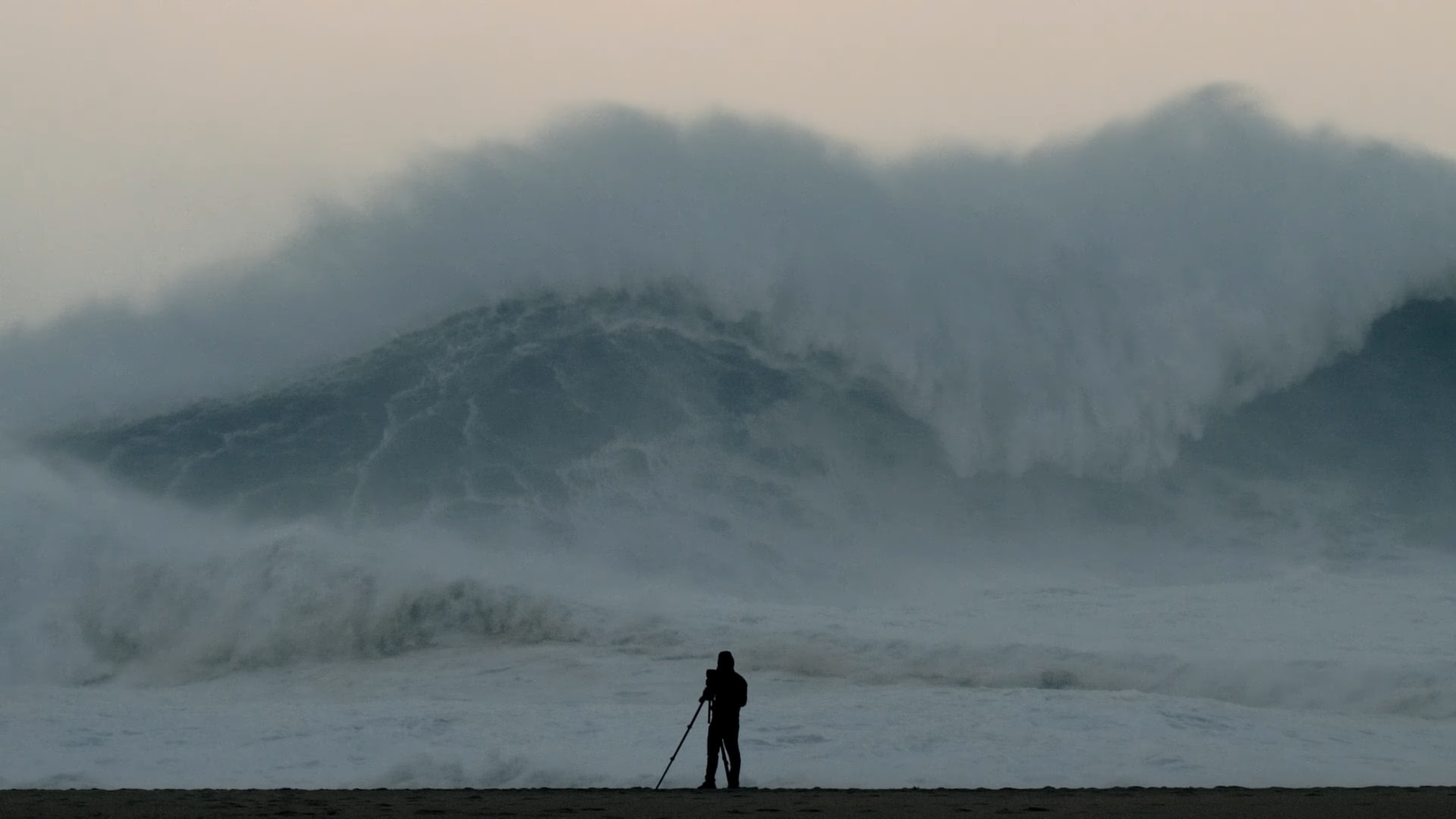 Ein Mensch mit einem Stativ steht am Strand vor riesigen, hohen Wellen im Meer.