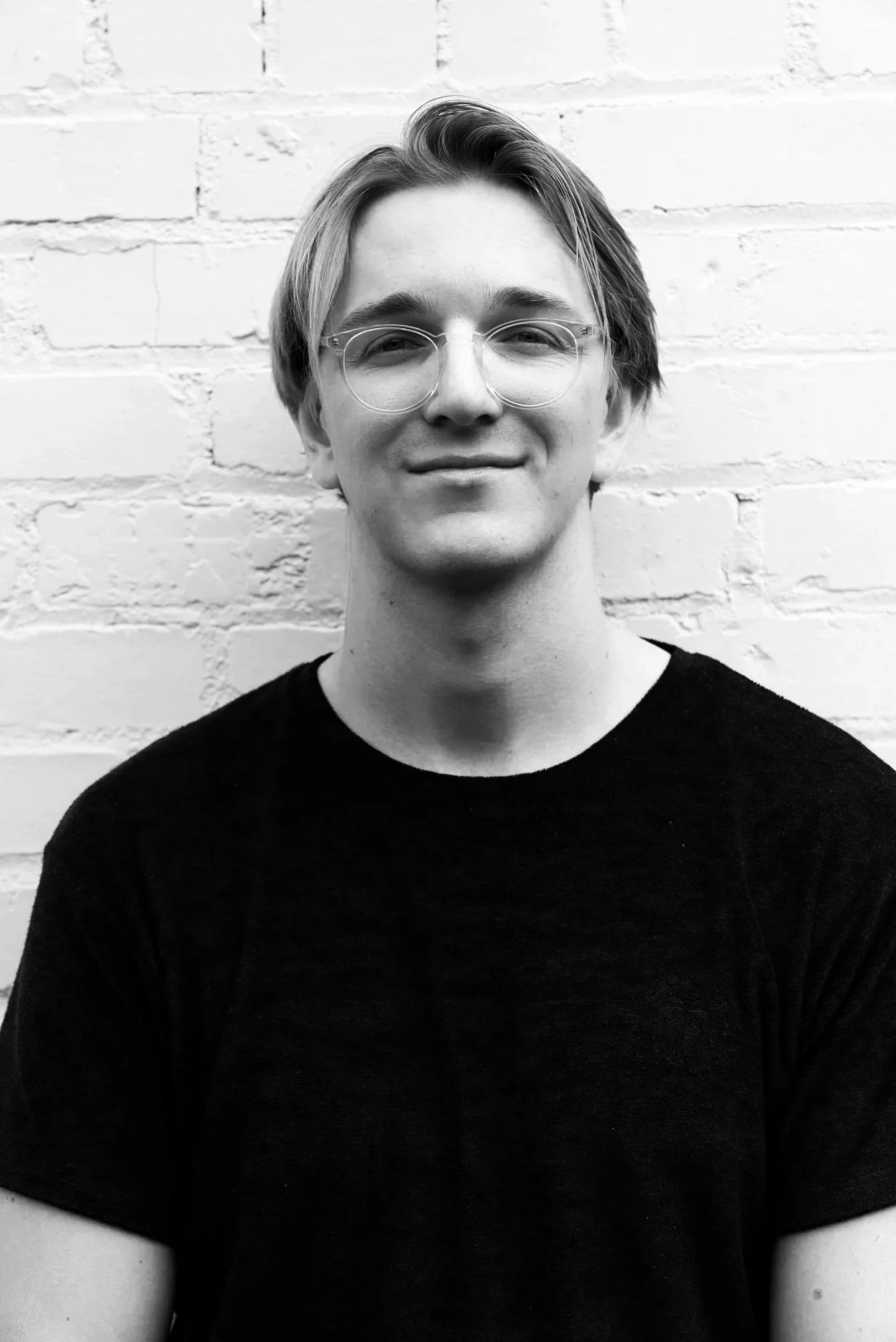 Black and white portrait of a young man with glasses, smiling, standing against a white brick wall.