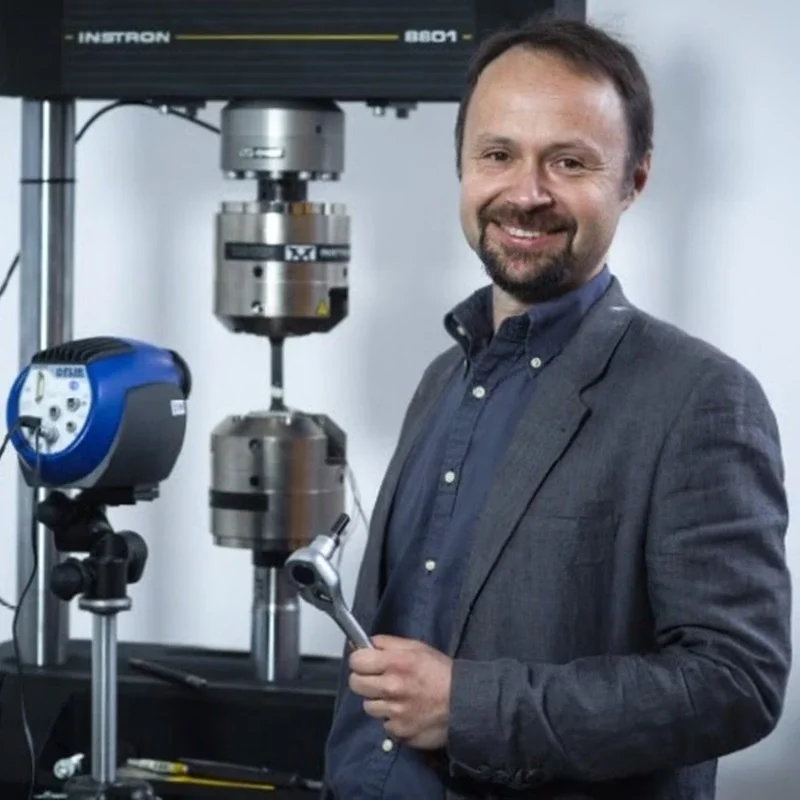 Man with a beard and dark hair in a gray blazer and navy blue shirt standing next to scientific equipment, holding a wrench.