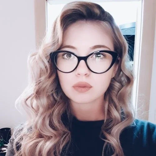 A woman with long wavy hair, wearing black glasses and a dark top, looking directly at the camera.