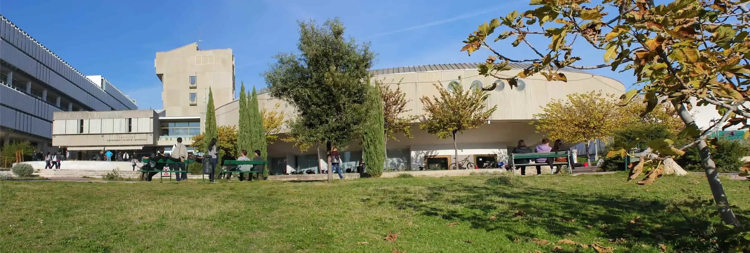Campus building with trees and benches, people sitting and walking outdoors on a sunny day.