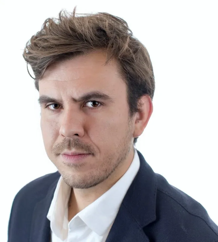 A man with tousled brown hair and light facial hair wearing a white dress shirt and dark blazer looking directly at the camera against a plain white background.