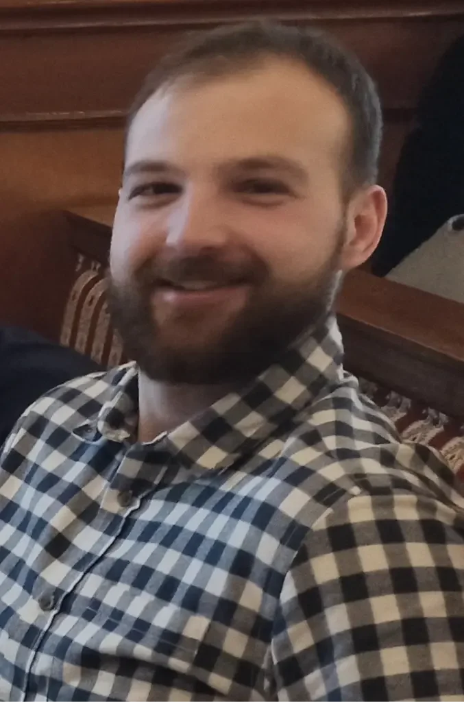 A smiling man with a beard, wearing a checkered shirt, sitting indoors with wooden furniture in the background.