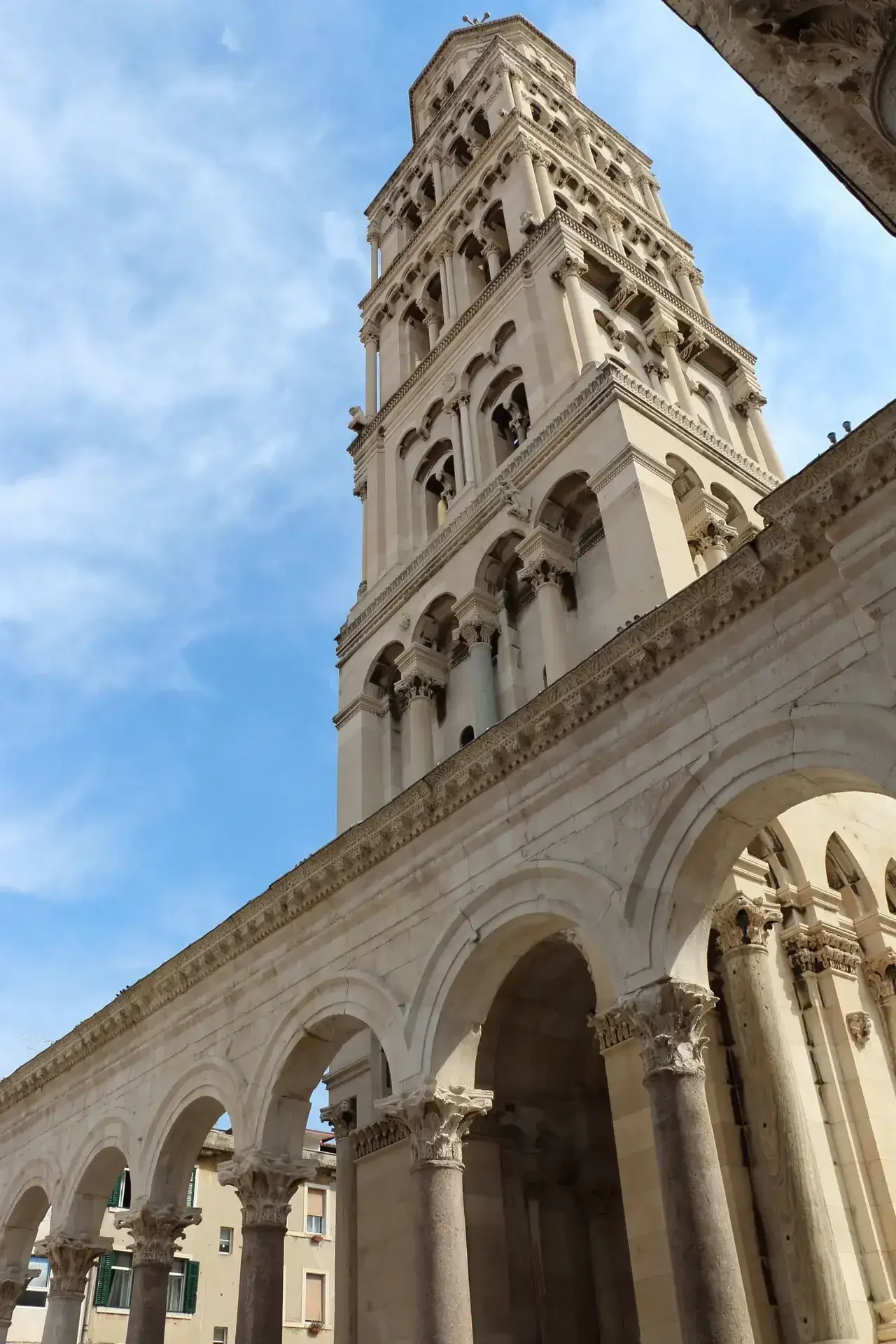 Low-angle view of an ornate historical tower with multiple arched levels, set against a blue sky with wispy clouds. Part of a building with classical architectural elements, including columns and arches, is visible at the bottom.