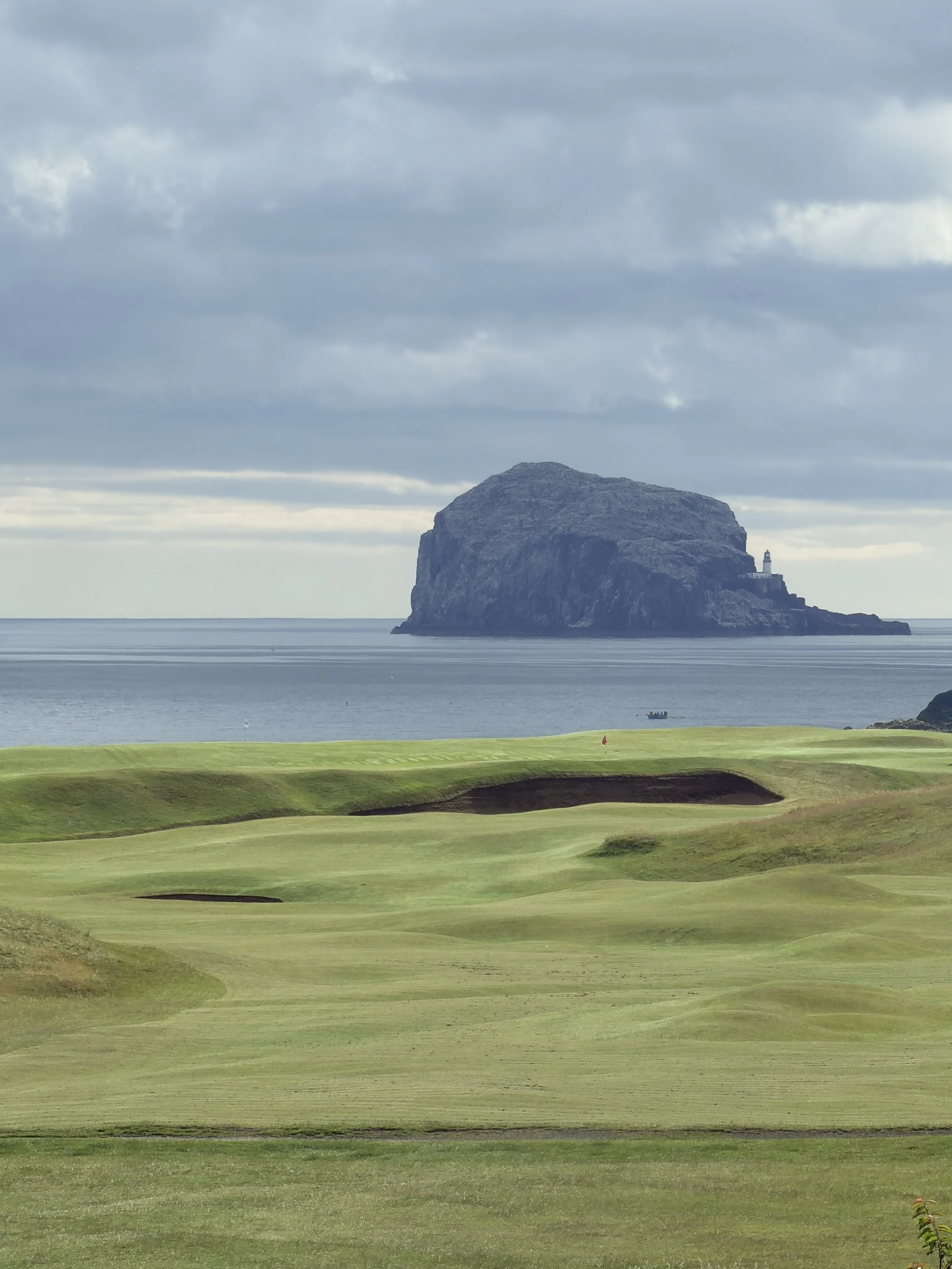 A scenic coastal view featuring a large rocky island with a lighthouse, the ocean, and a golf course with green grass and sand bunkers in the foreground, under a cloudy sky.