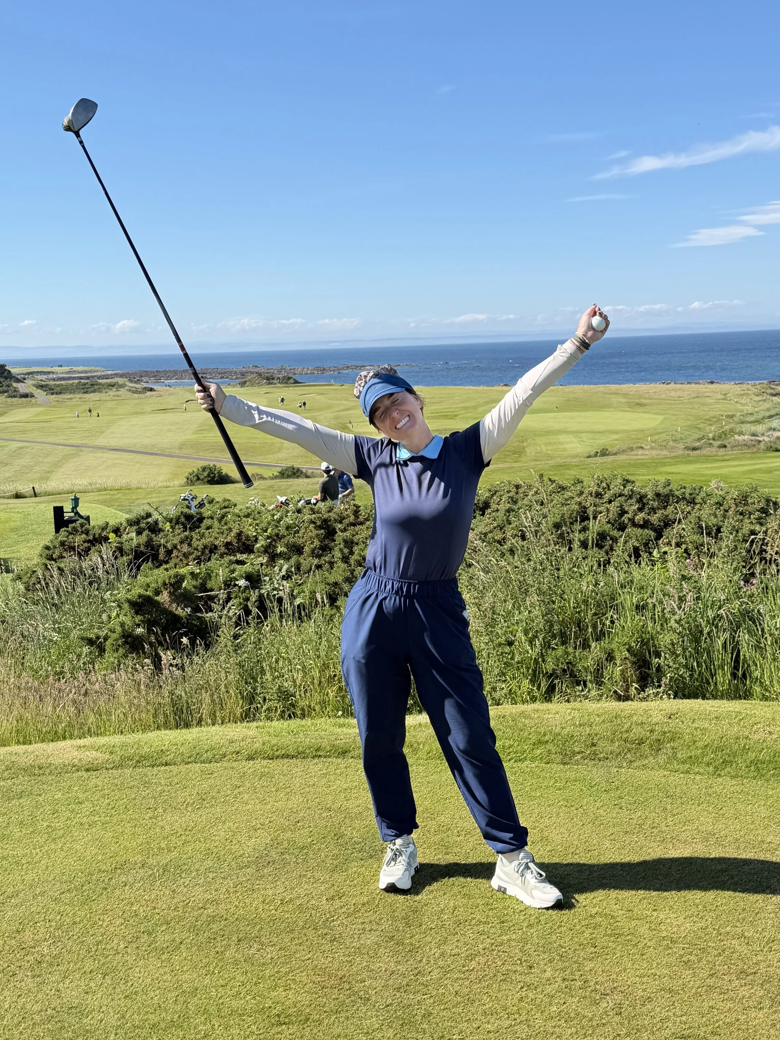 A woman standing on a golf course near the green, holding a golf club with arms outstretched, smiling, with ocean and blue sky in the background.