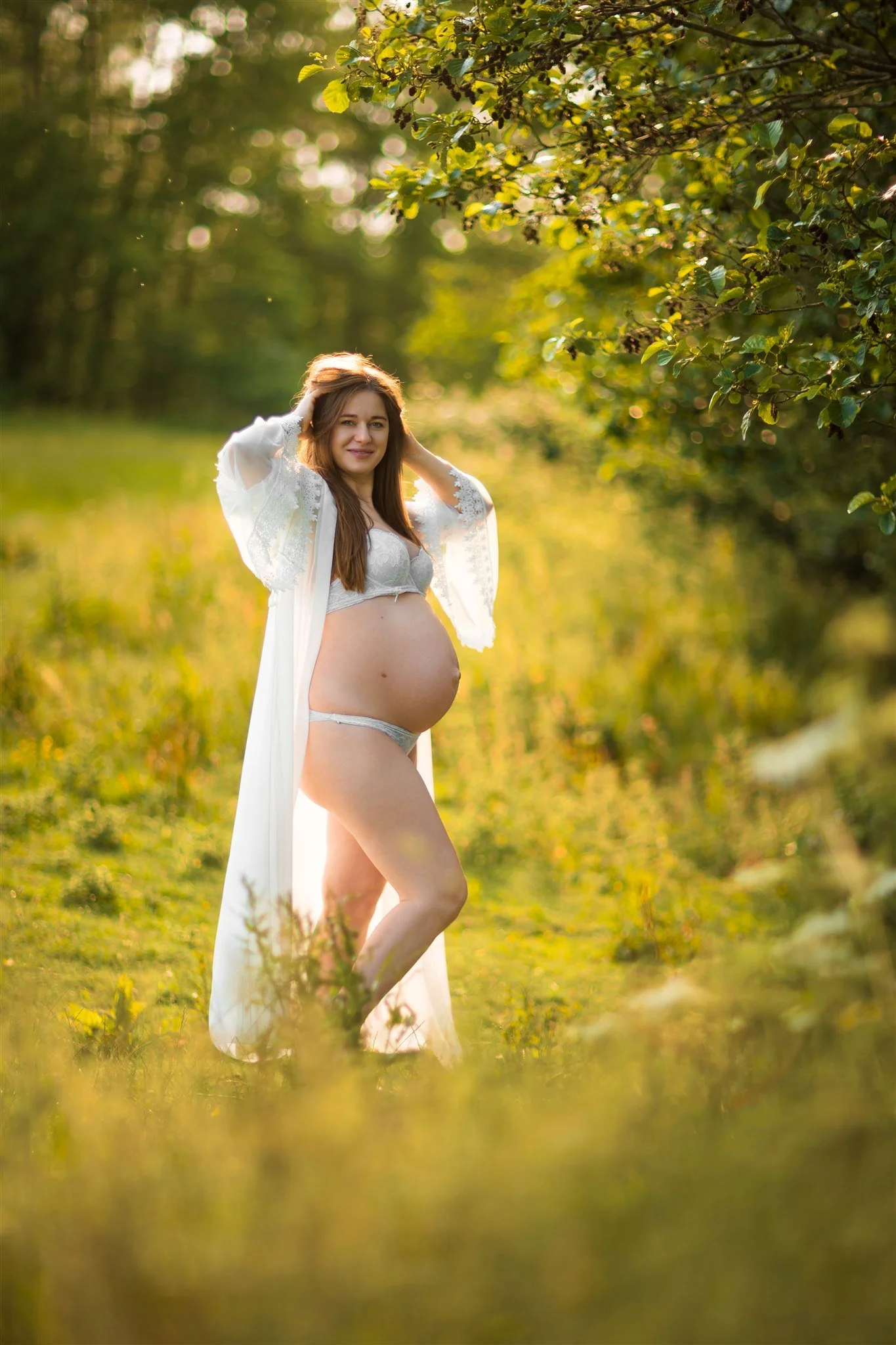 Pregnant woman in a white lace outfit stands in a sunny meadow, surrounded by greenery. She smiles gently, evoking a sense of peace and anticipation.