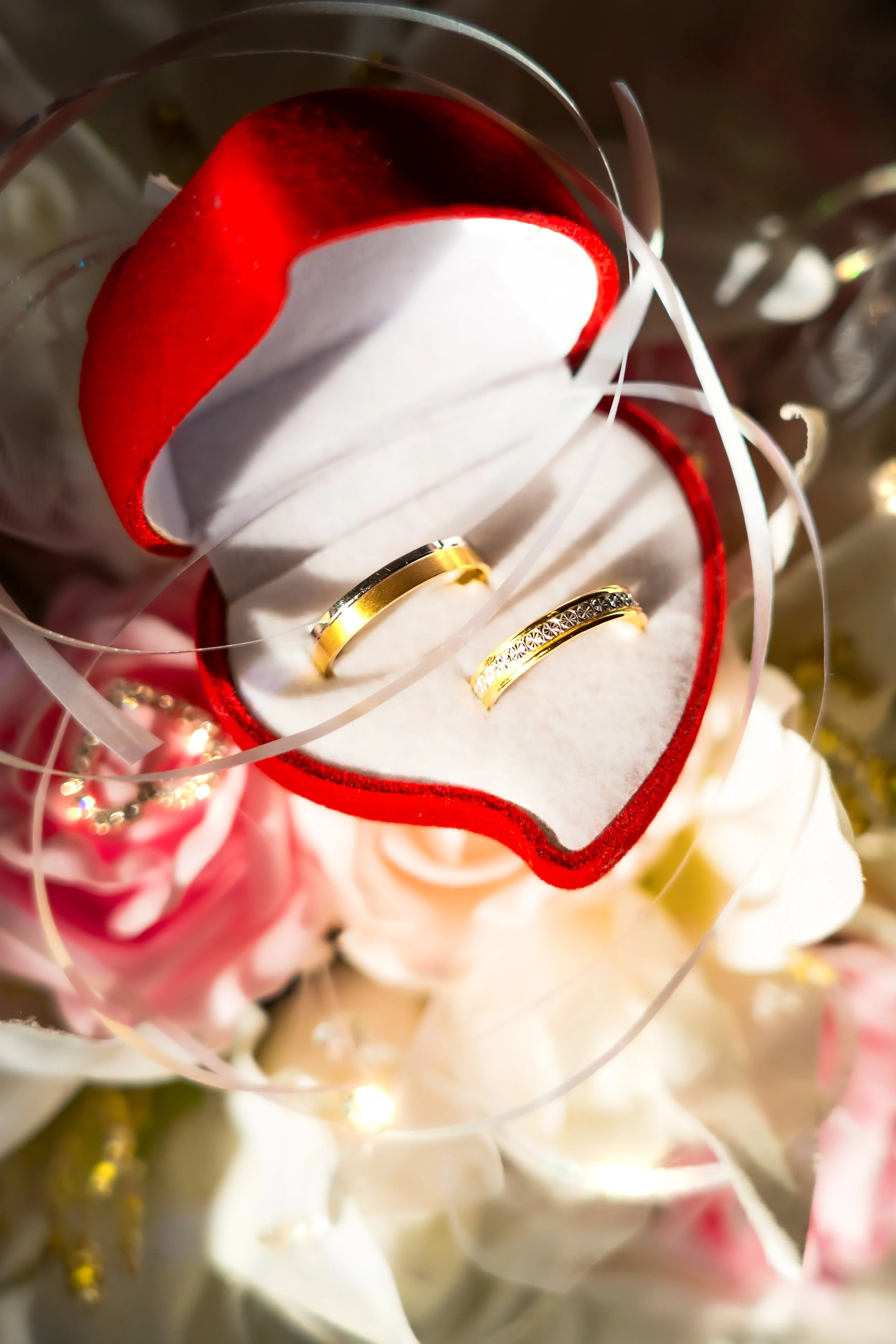 Two gold rings sit in a red heart-shaped velvet box, surrounded by delicate pink and white roses and ribbons, conveying romance and elegance.