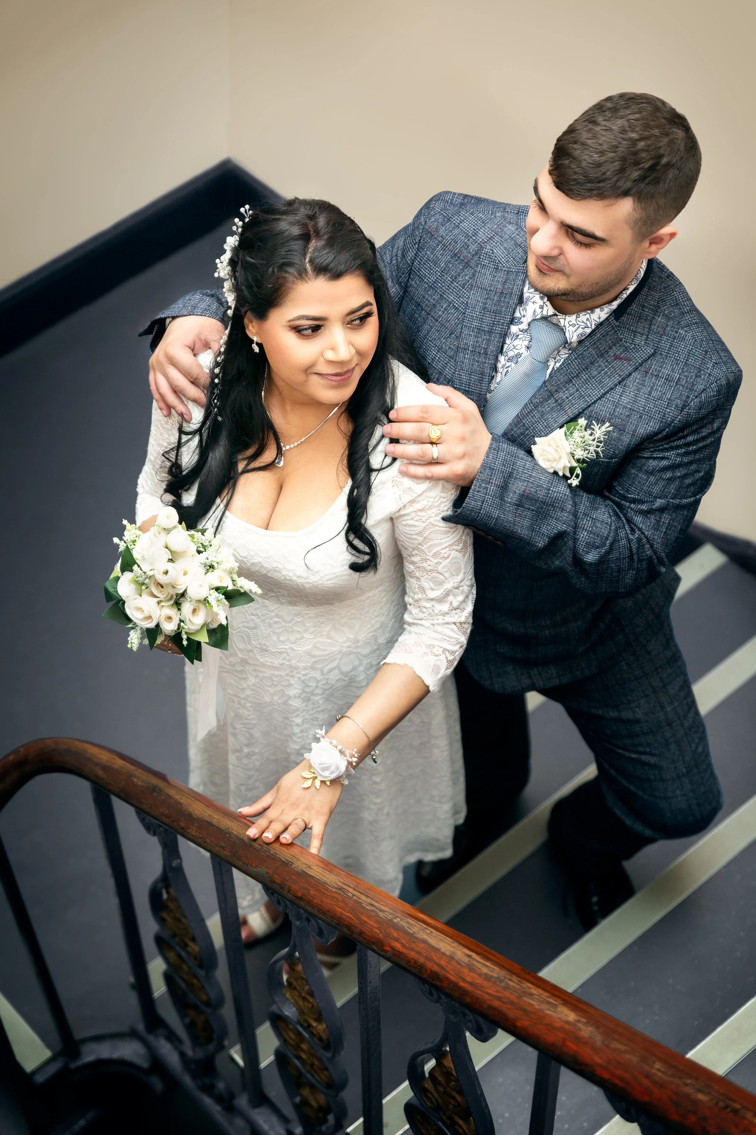 A couple in wedding attire poses on stairs: the woman in a lace dress holding white roses, and the man in a plaid suit. They share a joyful, tender moment.