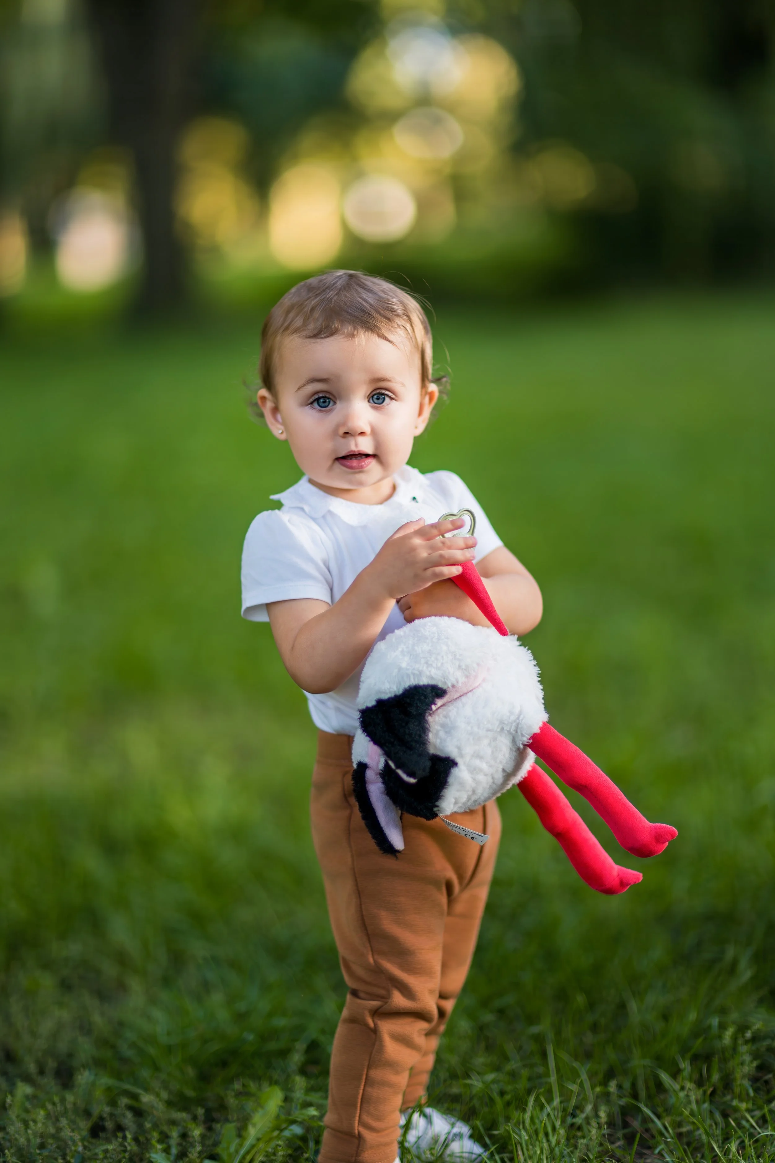 A toddler with short brown hair stands on green grass, holding a stuffed animal with red legs. The child wears a white shirt and brown pants, looking curious.