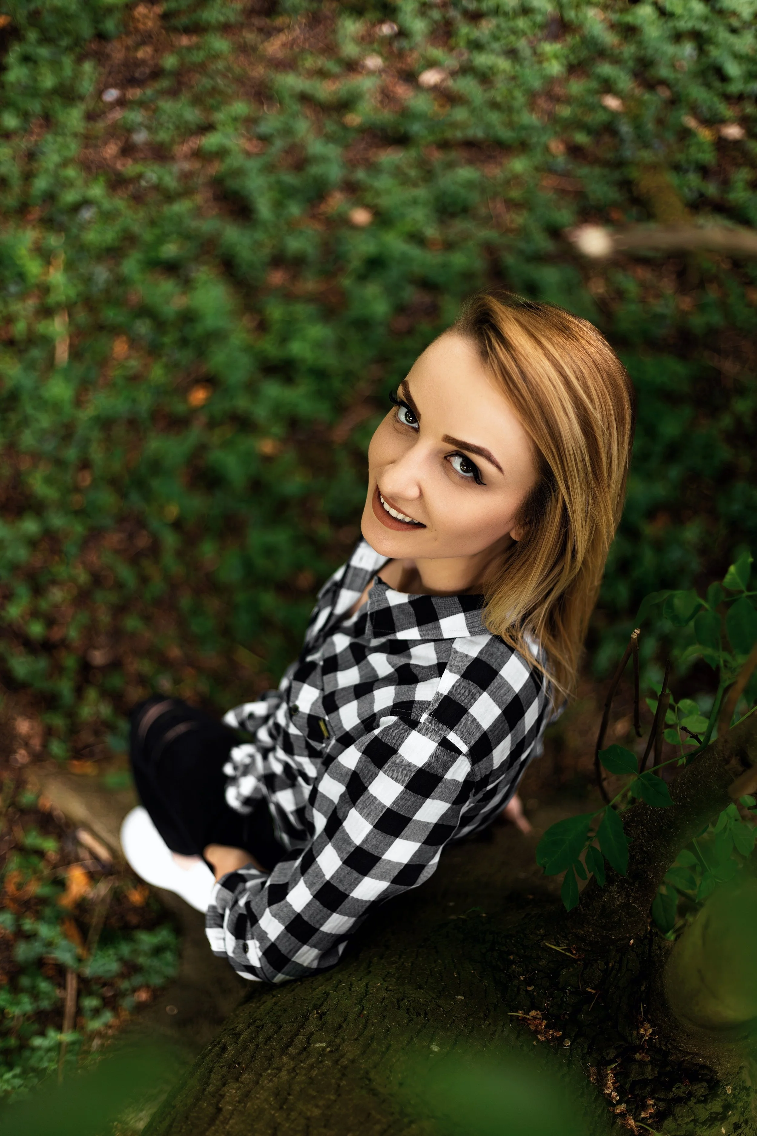 Portrait of a woman with dark blonde hair looking up at the camera while sitting at the bottom of a tree. She wears a black and white shirt, black jeans,  surrounded by lush green foliage, conveying and serene contemplative mood