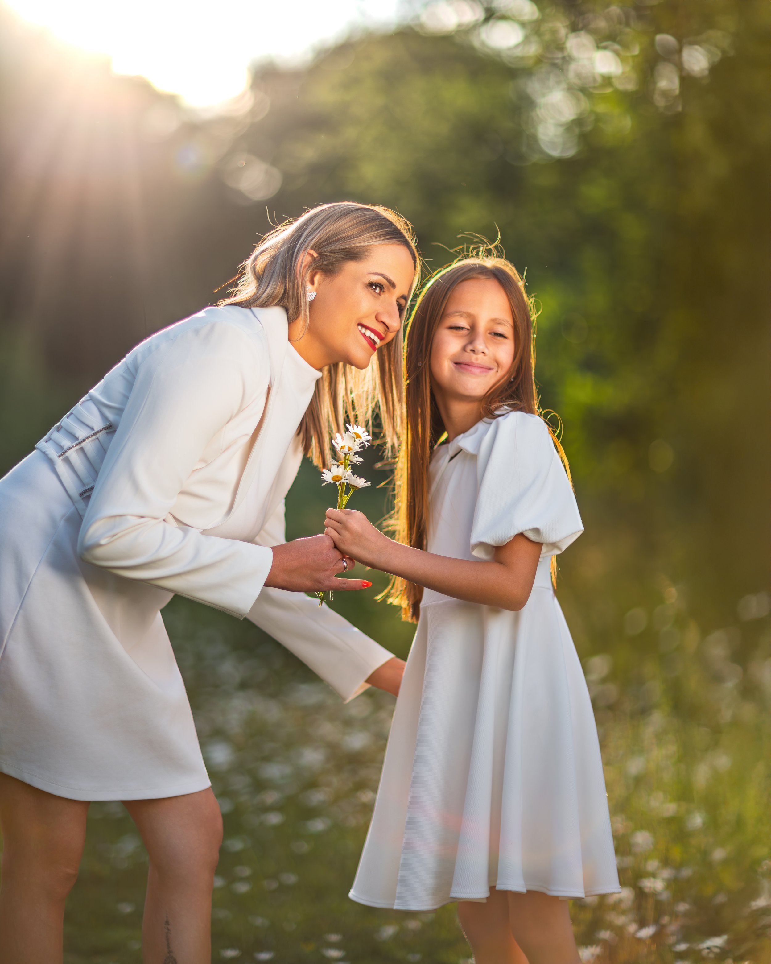 A woman and her young daughter, both in white dresses, stand in a sunlit park. The girl is giving daisies to her mother while they share a joyful moment, lush greenery all around them.