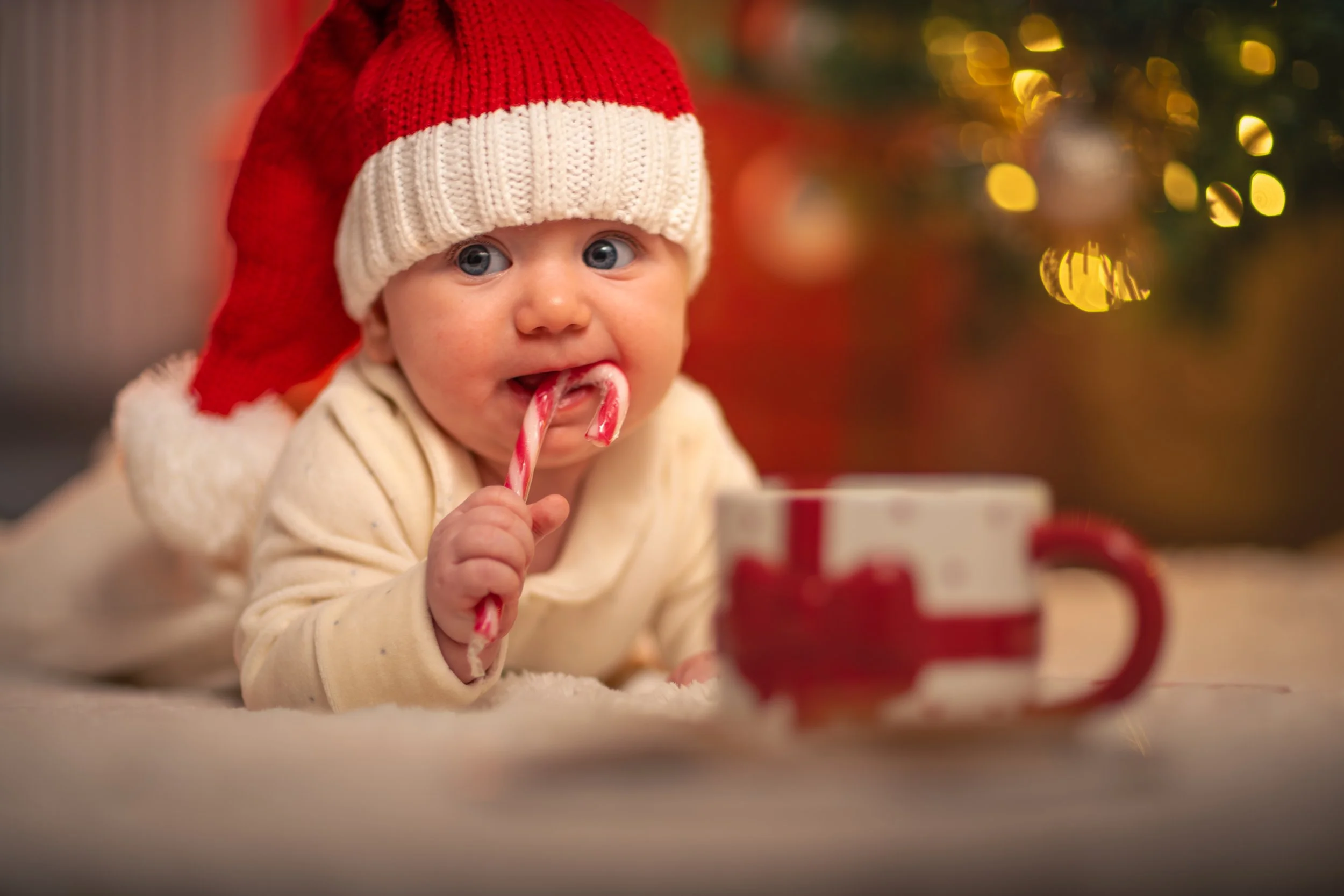 A baby wearing a red Santa hat and white outfit bites a candy cane, lying on a soft surface with a festive mug nearby. Background has warm, blurred holiday lights.