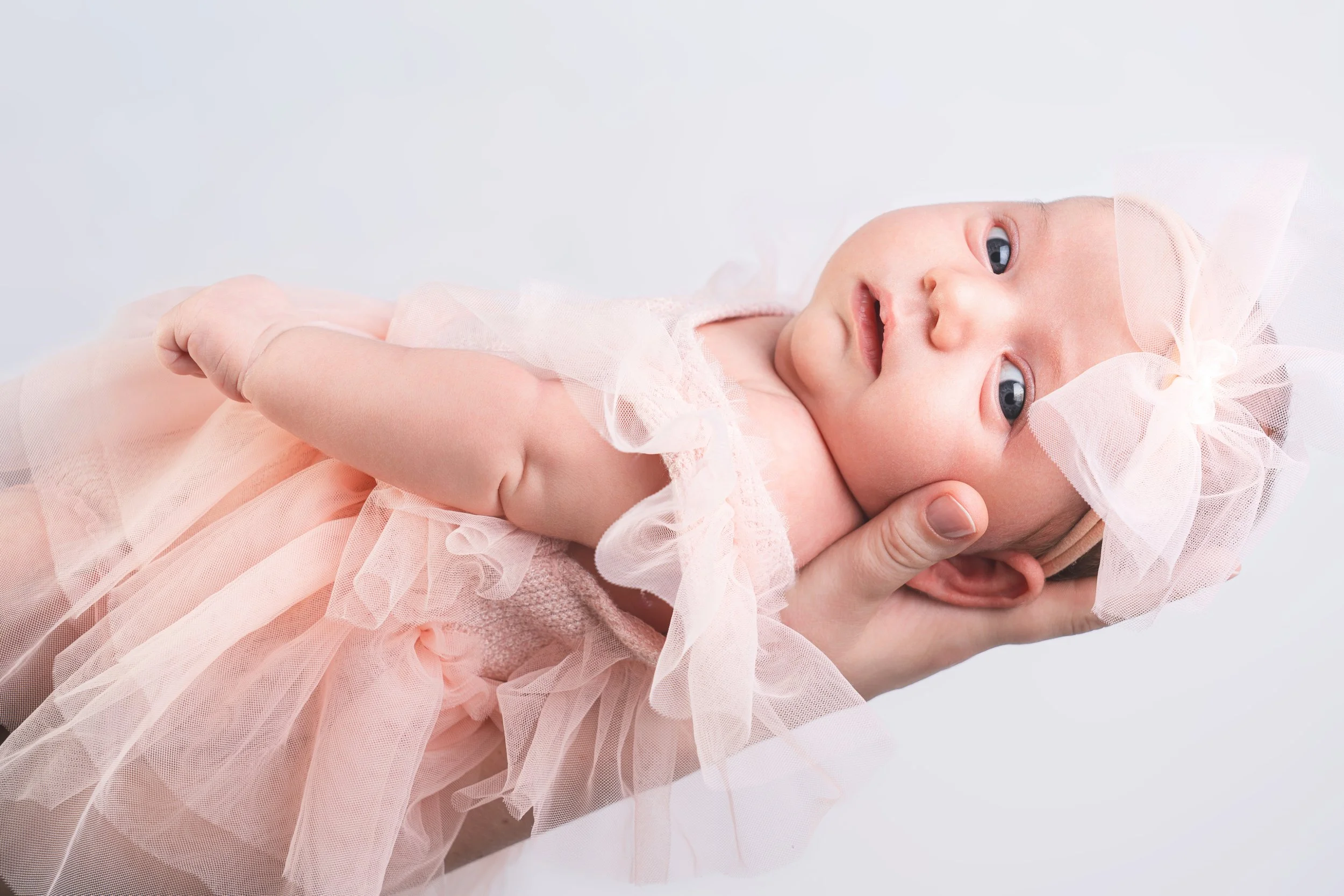A newborn baby is cradled in an adult's hands, wearing a delicate pink dress and headband. The baby has a calm expression against a soft, light background.