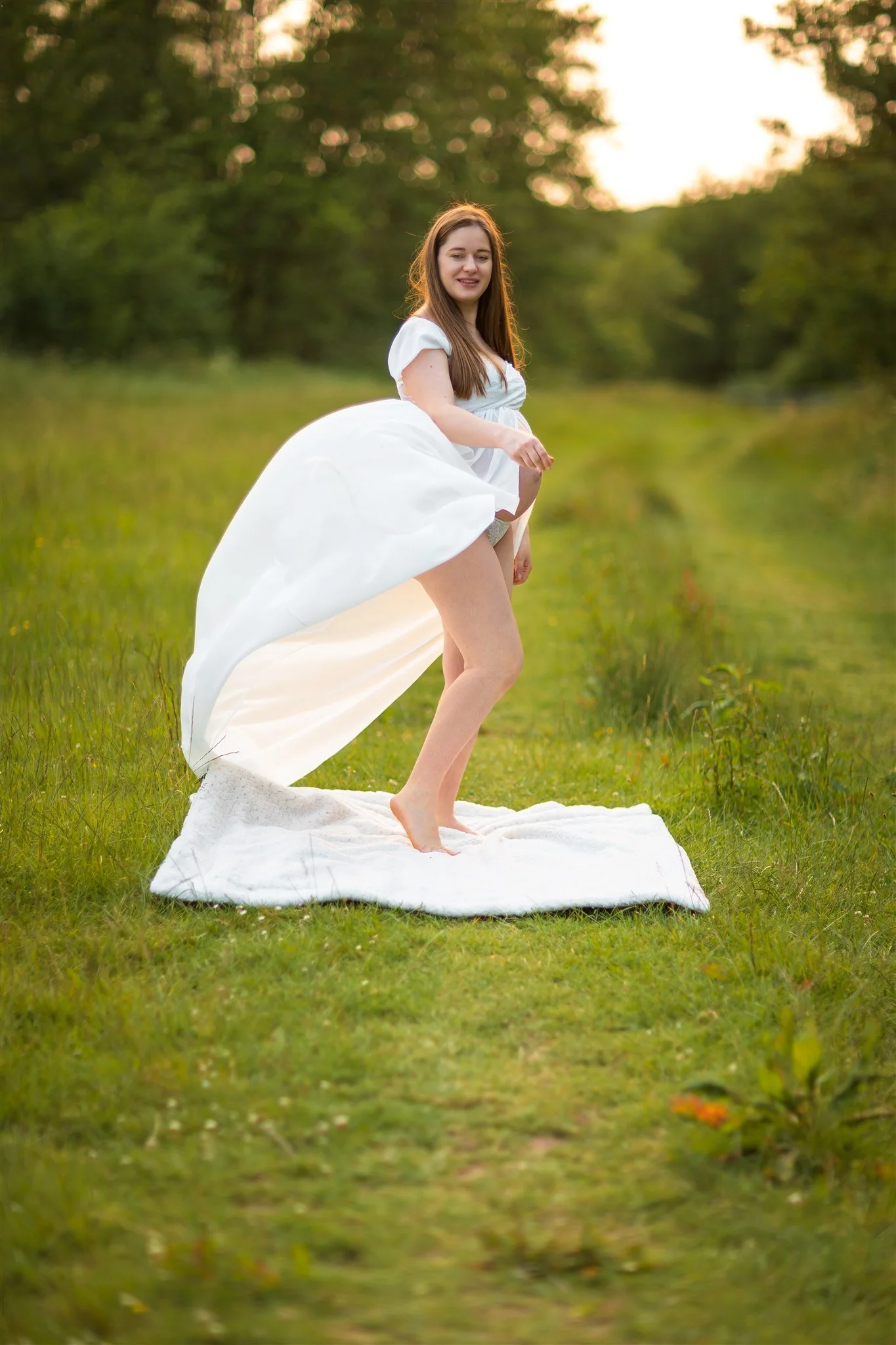A pregnant woman joyfully twirls on a white sheet in a lush green field, wearing a flowing white dress. The scene is serene, with soft golden light.
