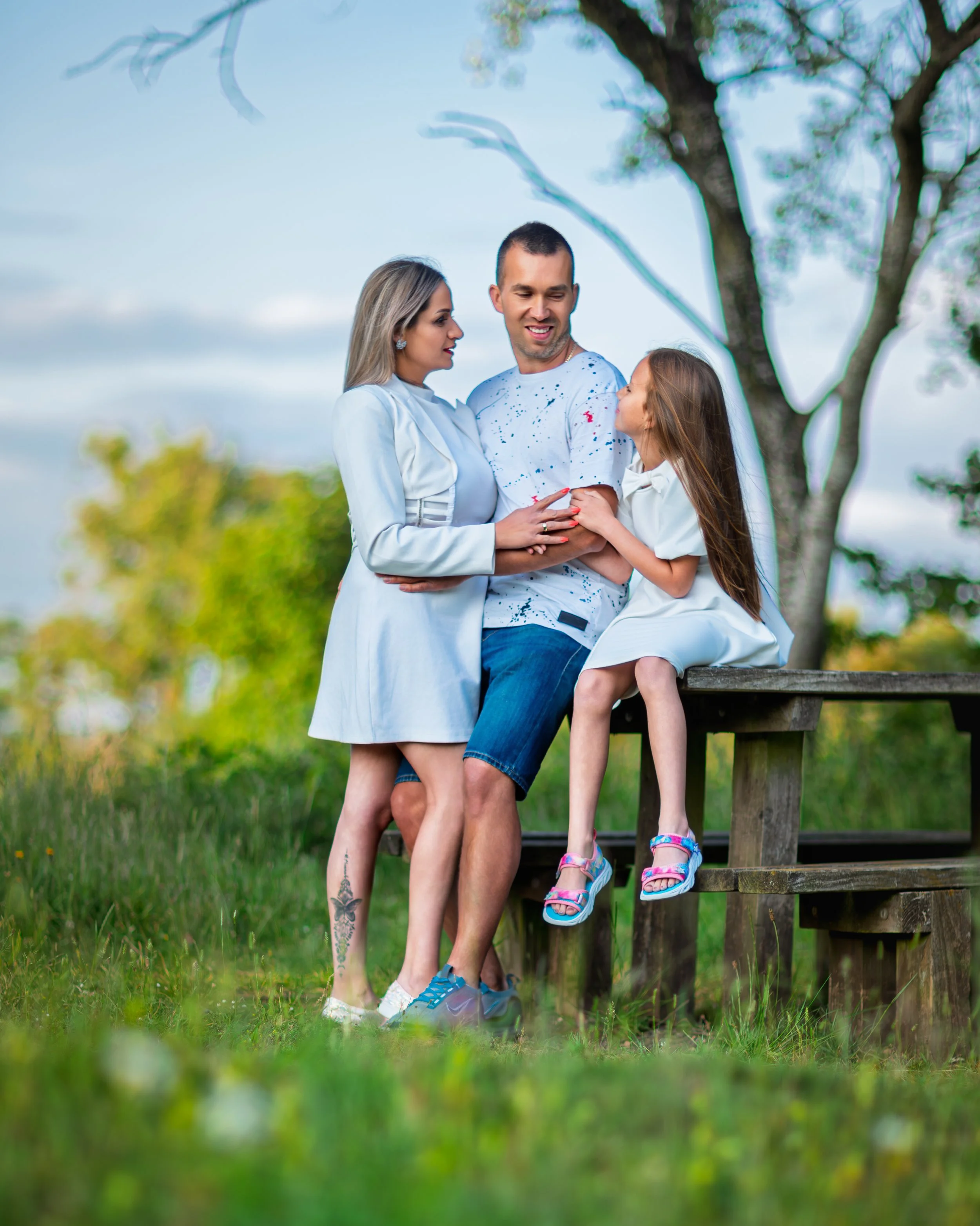 A smiling family poses outdoors on a picnic table, with greenery and trees in the background. The atmosphere is joyful and relaxed under a clear sky.