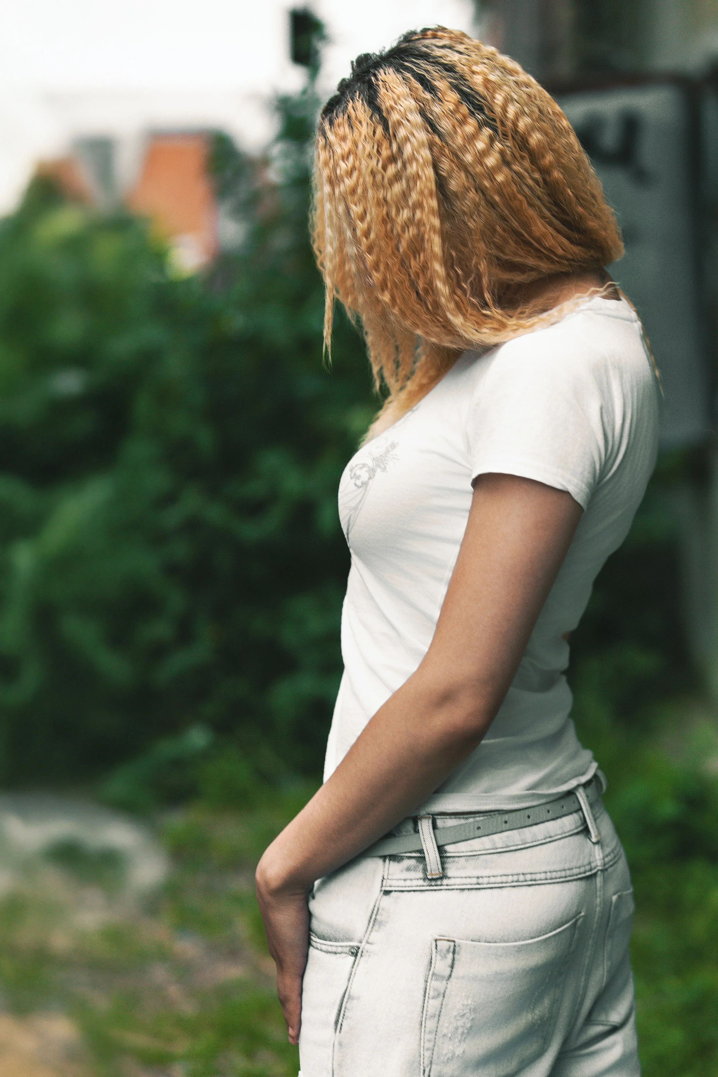 Faceless portrait of a blonde girl white t-shirt and jeans in front of a bush in an industrial area