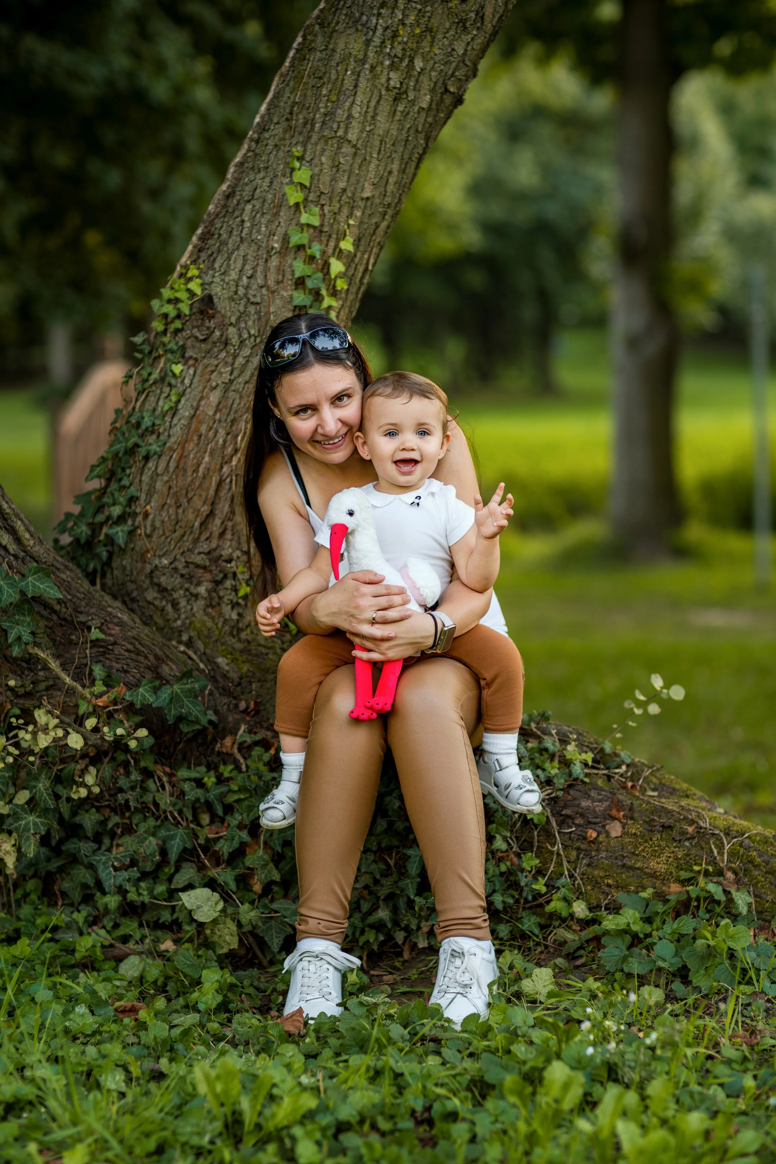 A woman sits under a large tree in a park, holding a smiling baby on her lap. Both are wearing casual clothes. The scene conveys joy and warmth.