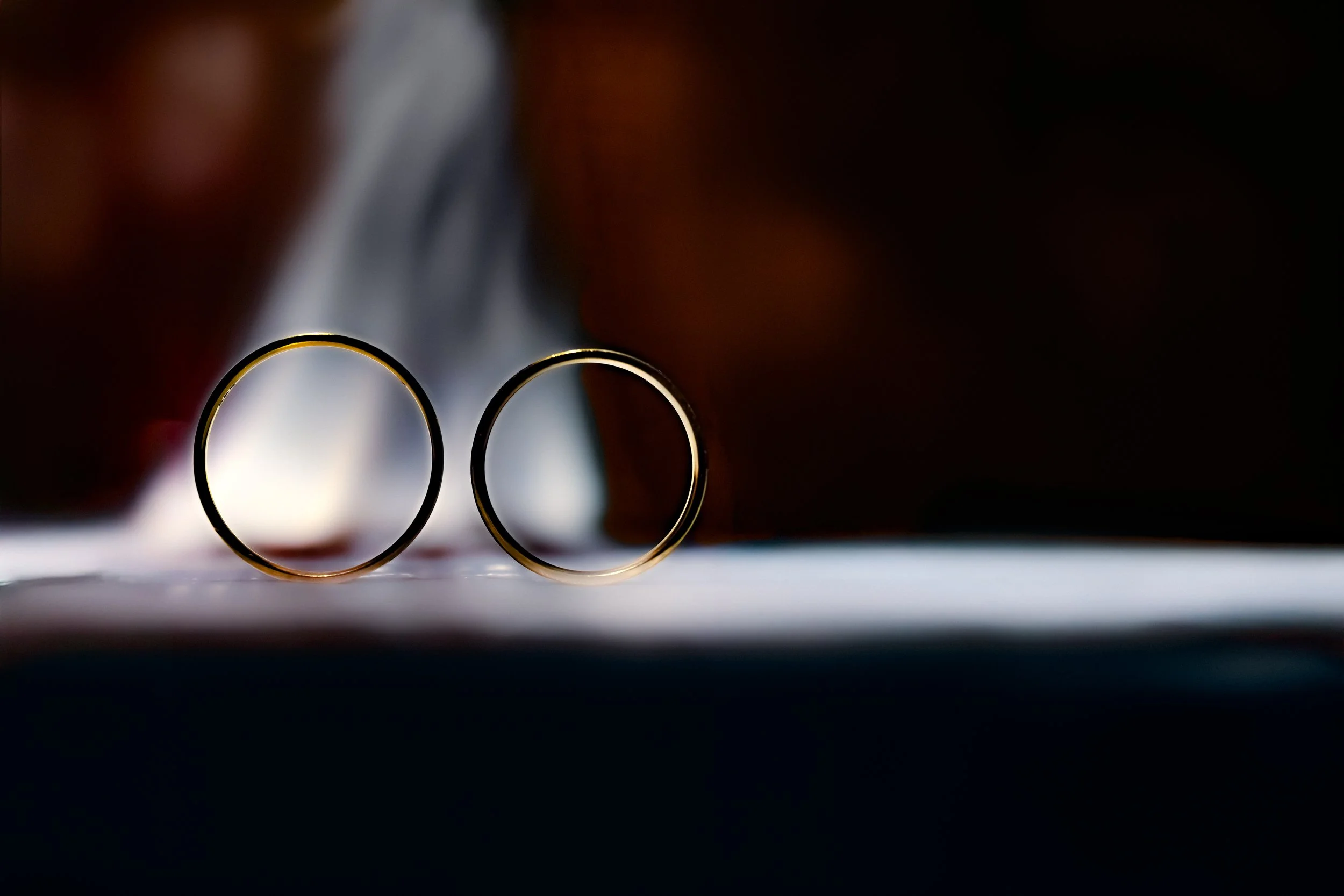 Two gold rings stand upright on a surface, backlit by a soft, blurred glow. The scene conveys a sense of elegance and intimacy.