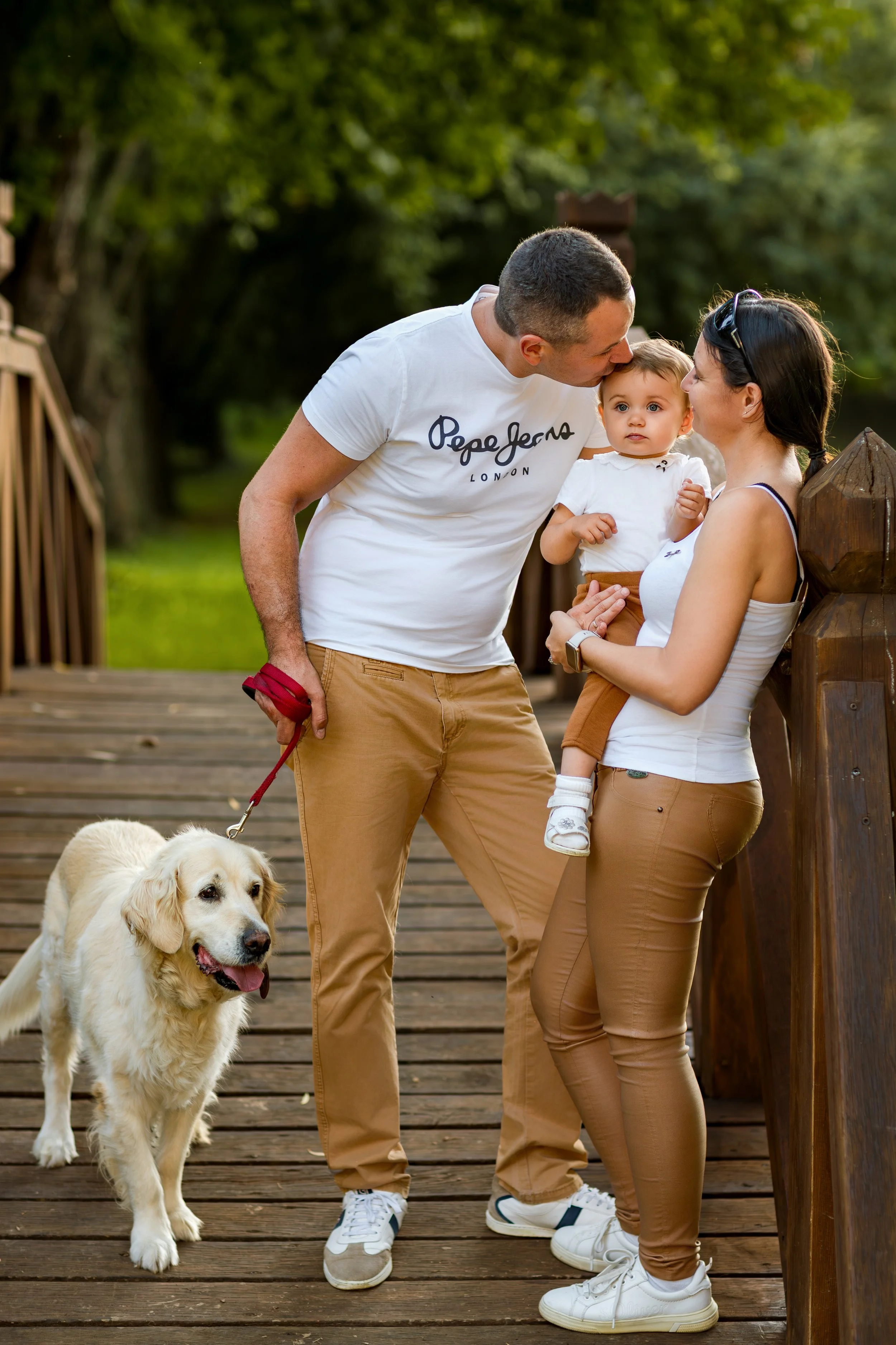 A family stands on a wooden bridge in a park. A man leans in to kiss a woman holding a baby. A happy golden retriever is beside them.