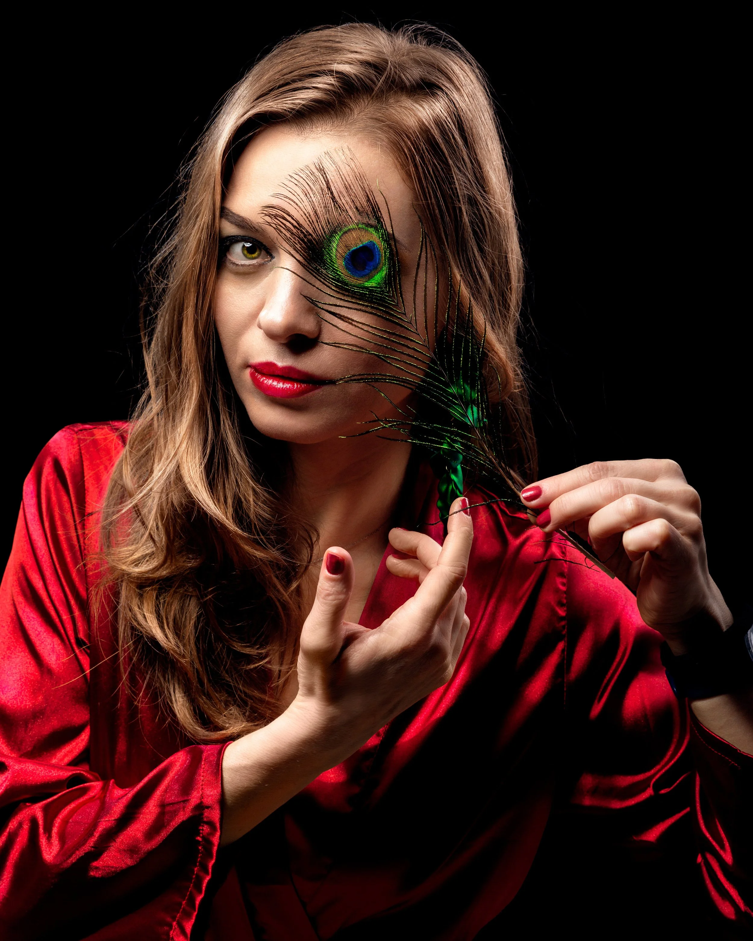 Woman in red silk blouse holds a peacock feather over her eye against a dark background. Her expression is thoughtful, creating a mysterious tone.