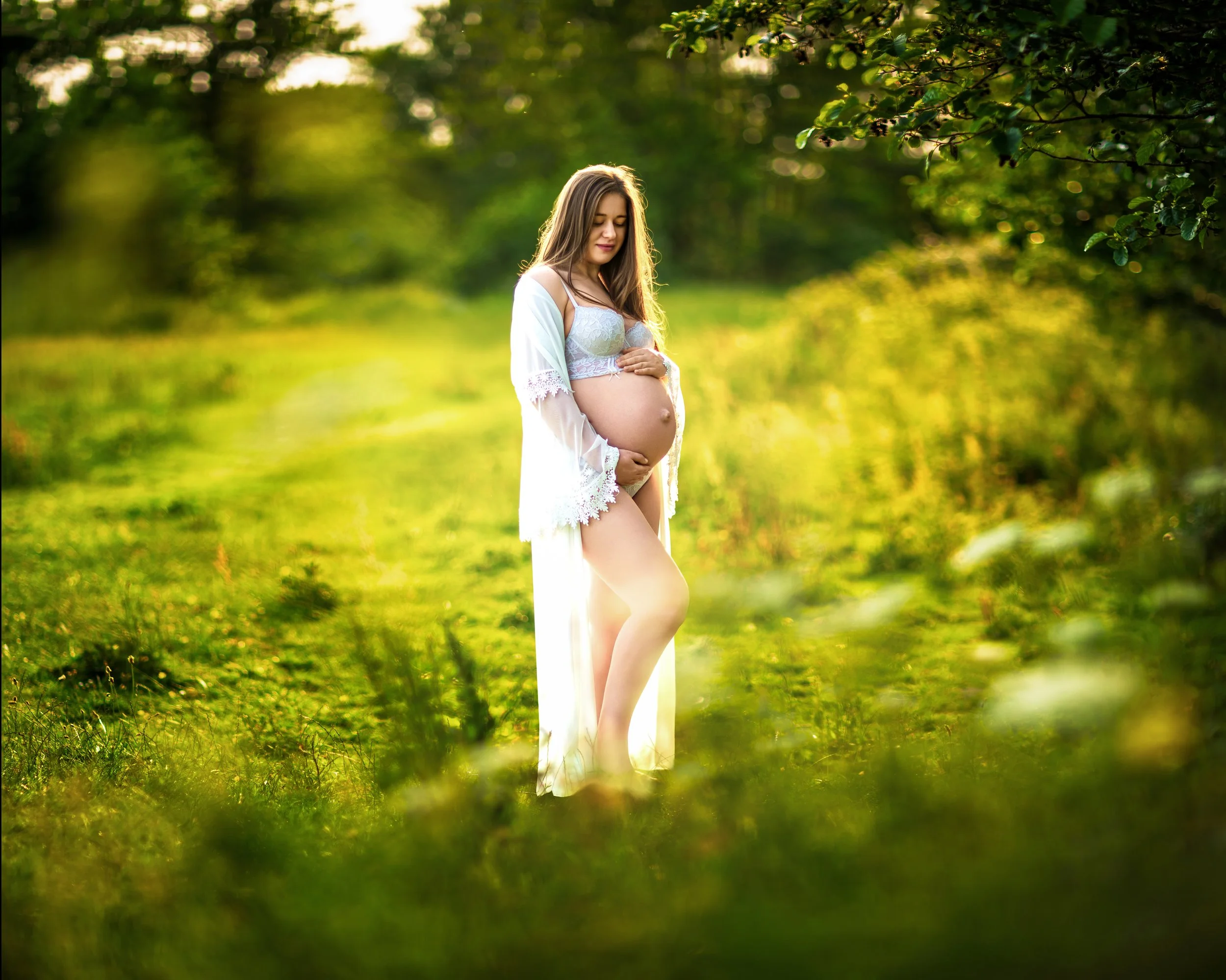 A pregnant woman in a flowing white dress stands in a serene, sunlit meadow, gently cradling her belly. The scene conveys peace and anticipation.