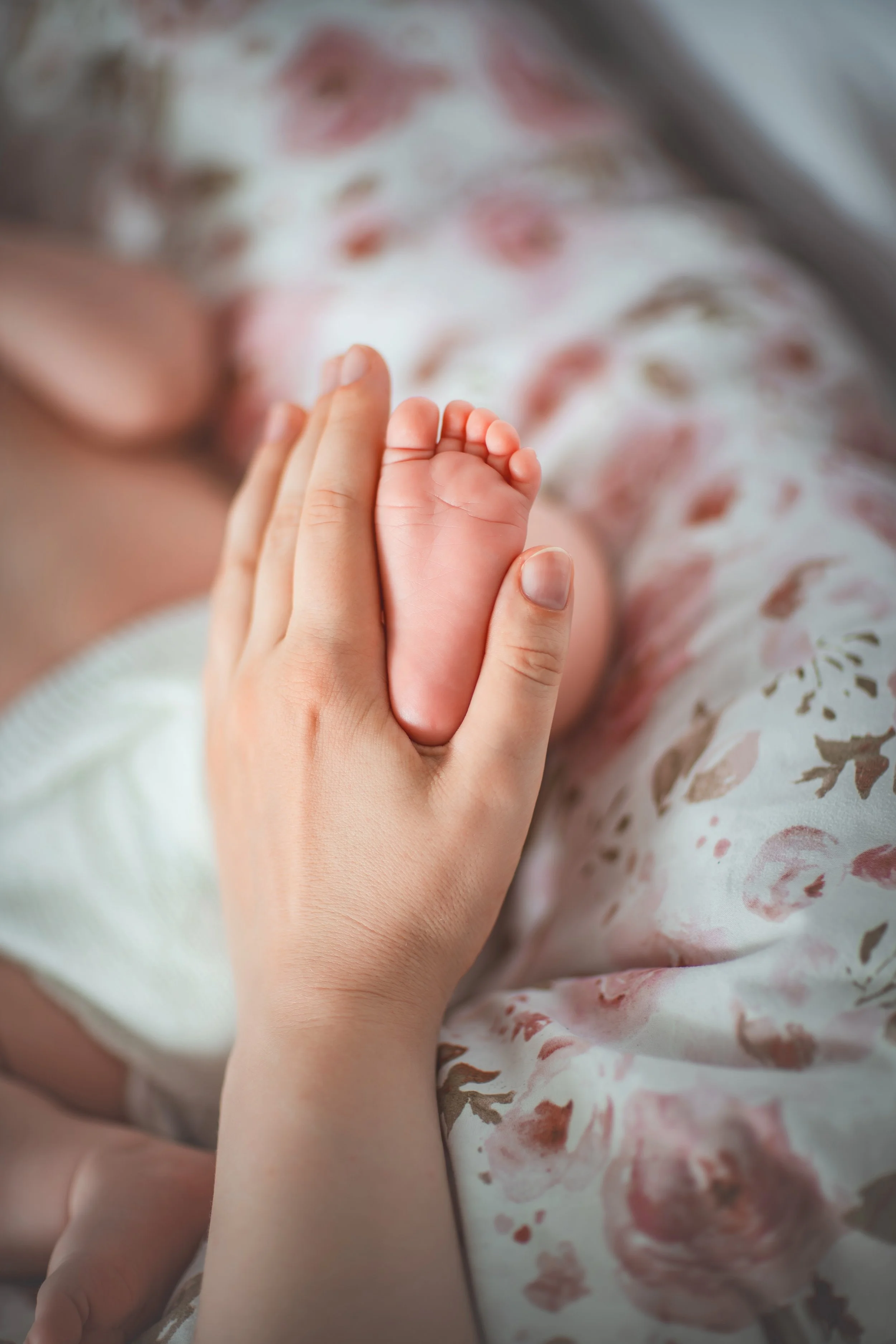 A gentle adult hand cradles a tiny baby foot on a floral patterned blanket, evoking warmth and tenderness. The scene conveys love and protection.
