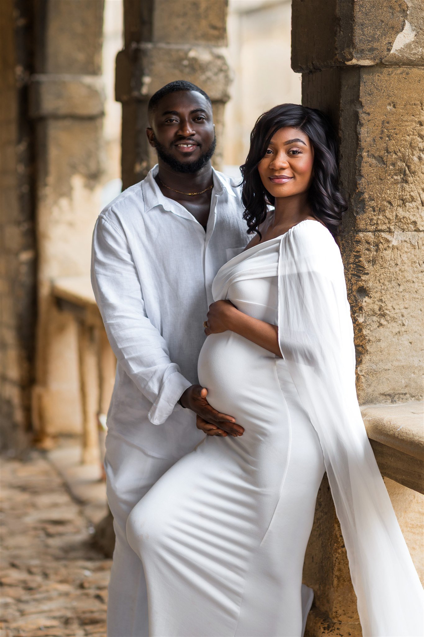 A couple stands in an ancient stone corridor. The woman, in a flowing white dress, cradles her baby bump, while the man stands beside her, smiling warmly.
