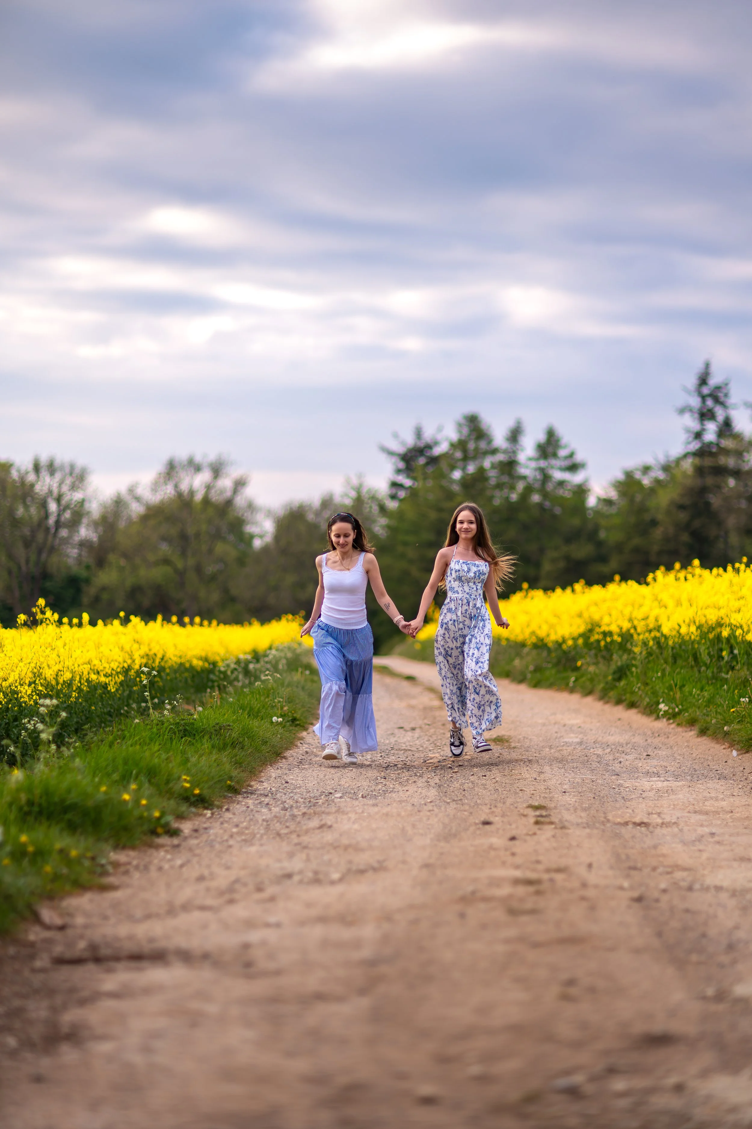 A woman and her niece walking hand in hand along a countryside path beside blooming yellow fields under a cloudy spring sky.