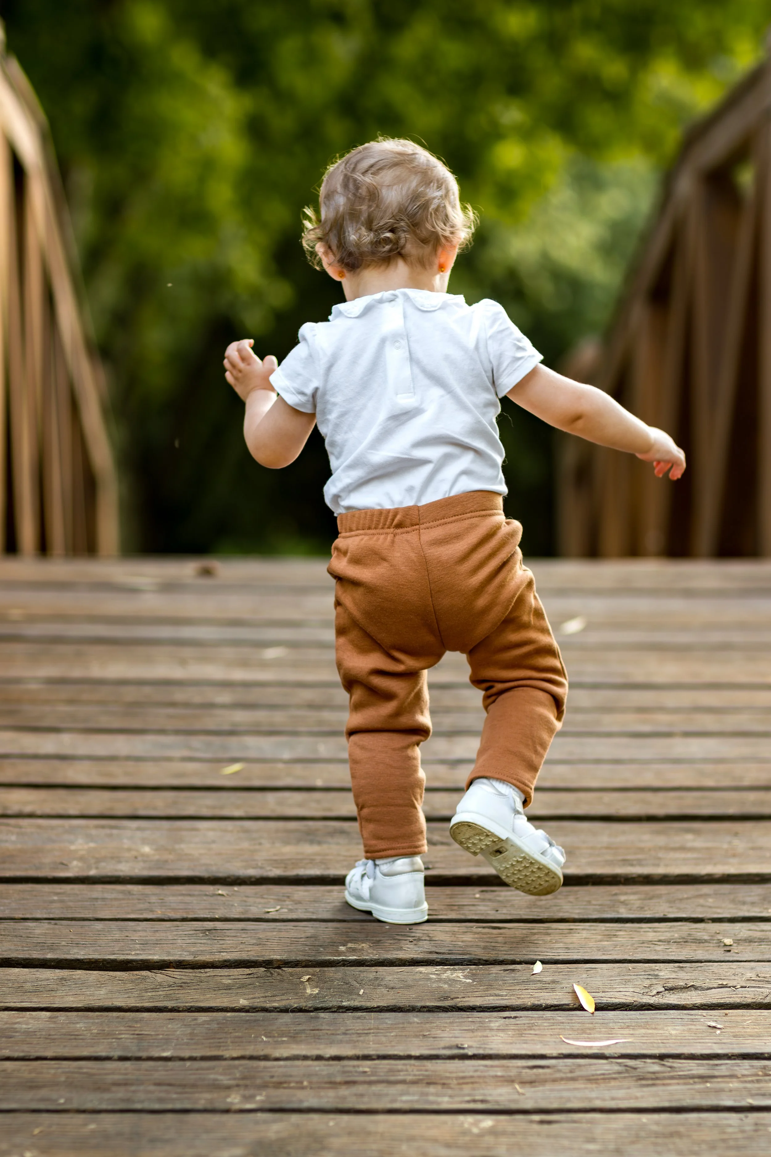 A toddler in a white shirt and brown pants confidently walks on a wooden bridge, surrounded by lush greenery, evoking a sense of exploration and joy.