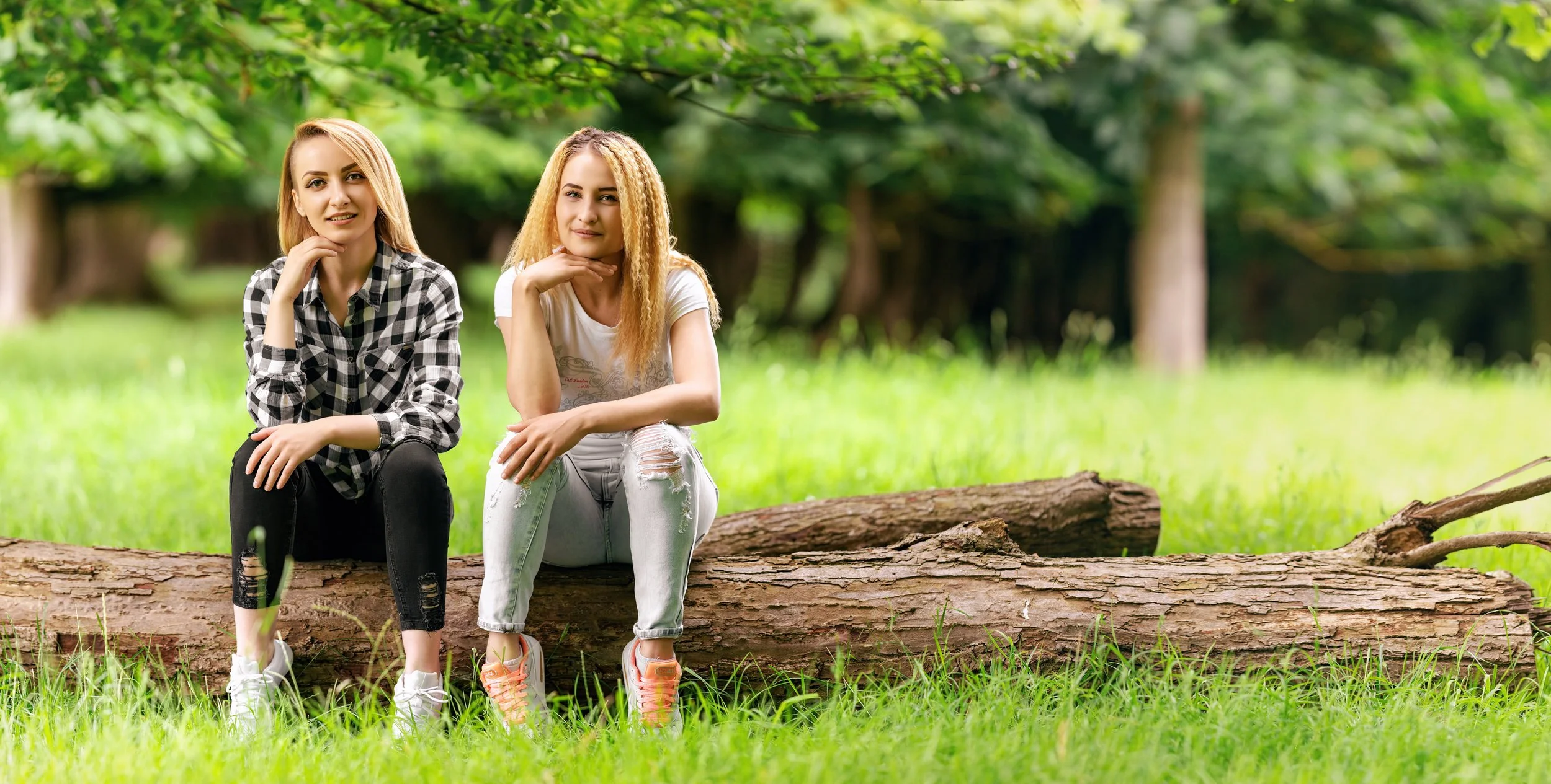 Portrait of  two women sit on a log in a lush  green park, smiling and relaxed. One wears a shirt the other wears a white T-shirt. Trees fall out of focus in the background 