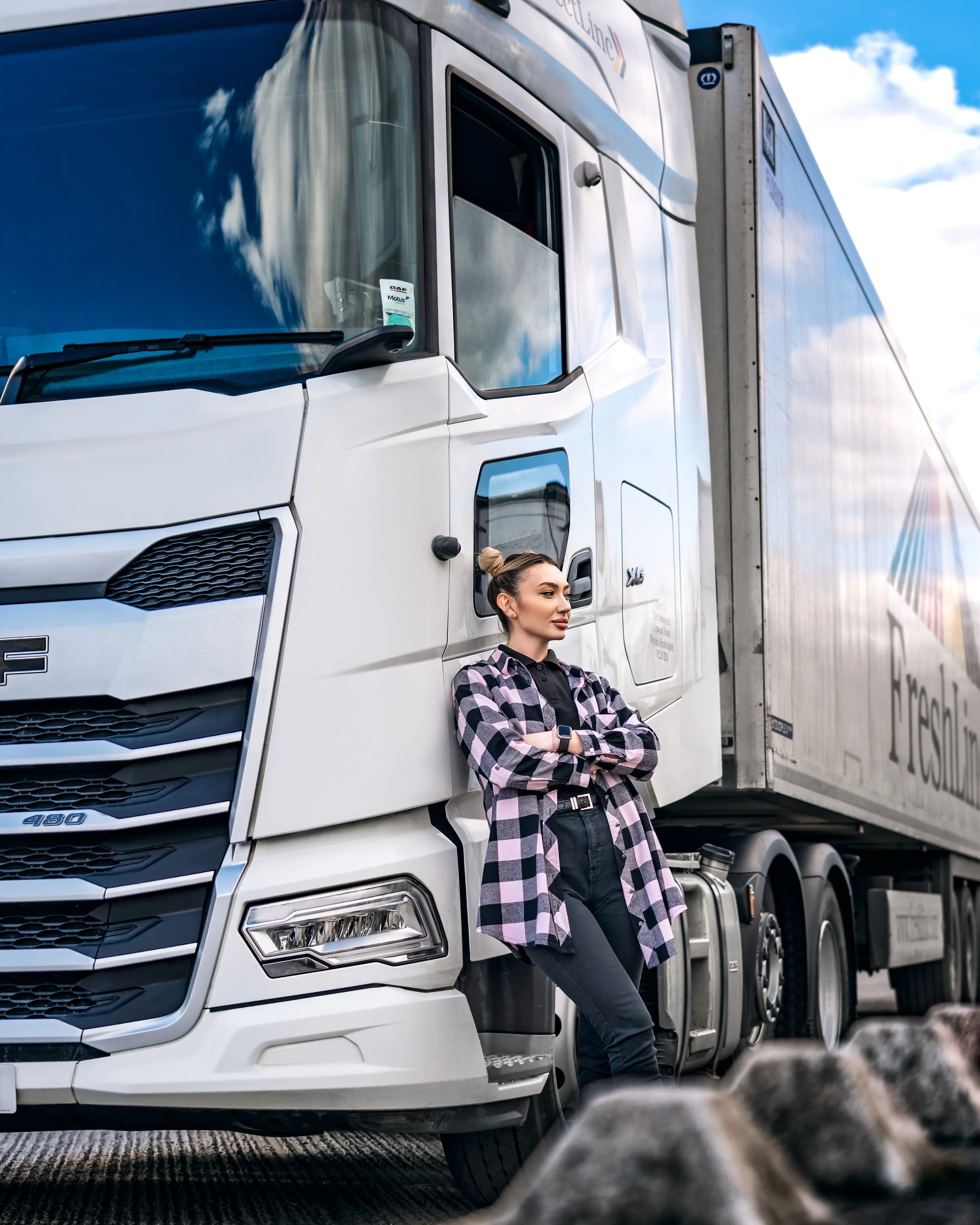 Portrait of a young woman leans confidently against a large white DAF lorry under a partly cloudy blue sky. They wear a pink checked shirt and black jeans