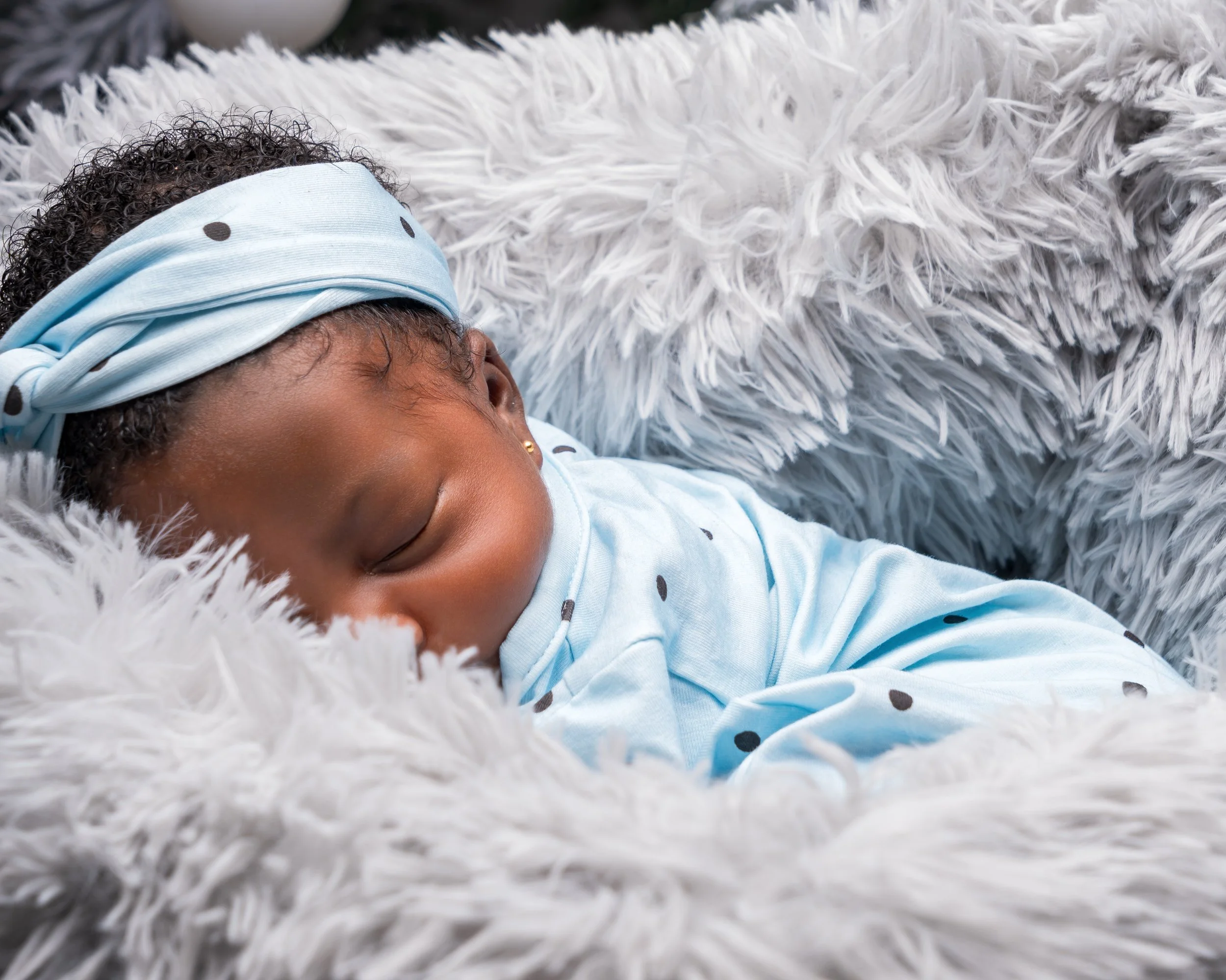 A sleeping baby wears a light blue outfit with black polka dots, resting on a fluffy gray blanket. The scene conveys serenity and warmth.