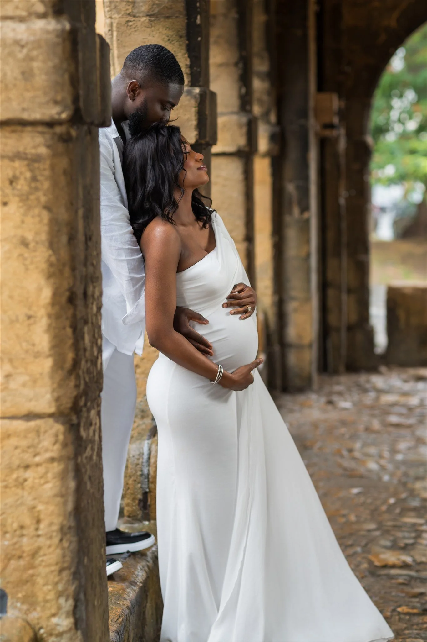 A couple embraces lovingly under a stone archway. The pregnant woman in a white dress smiles upward, while the man leans close, creating an intimate, joyful scene.
