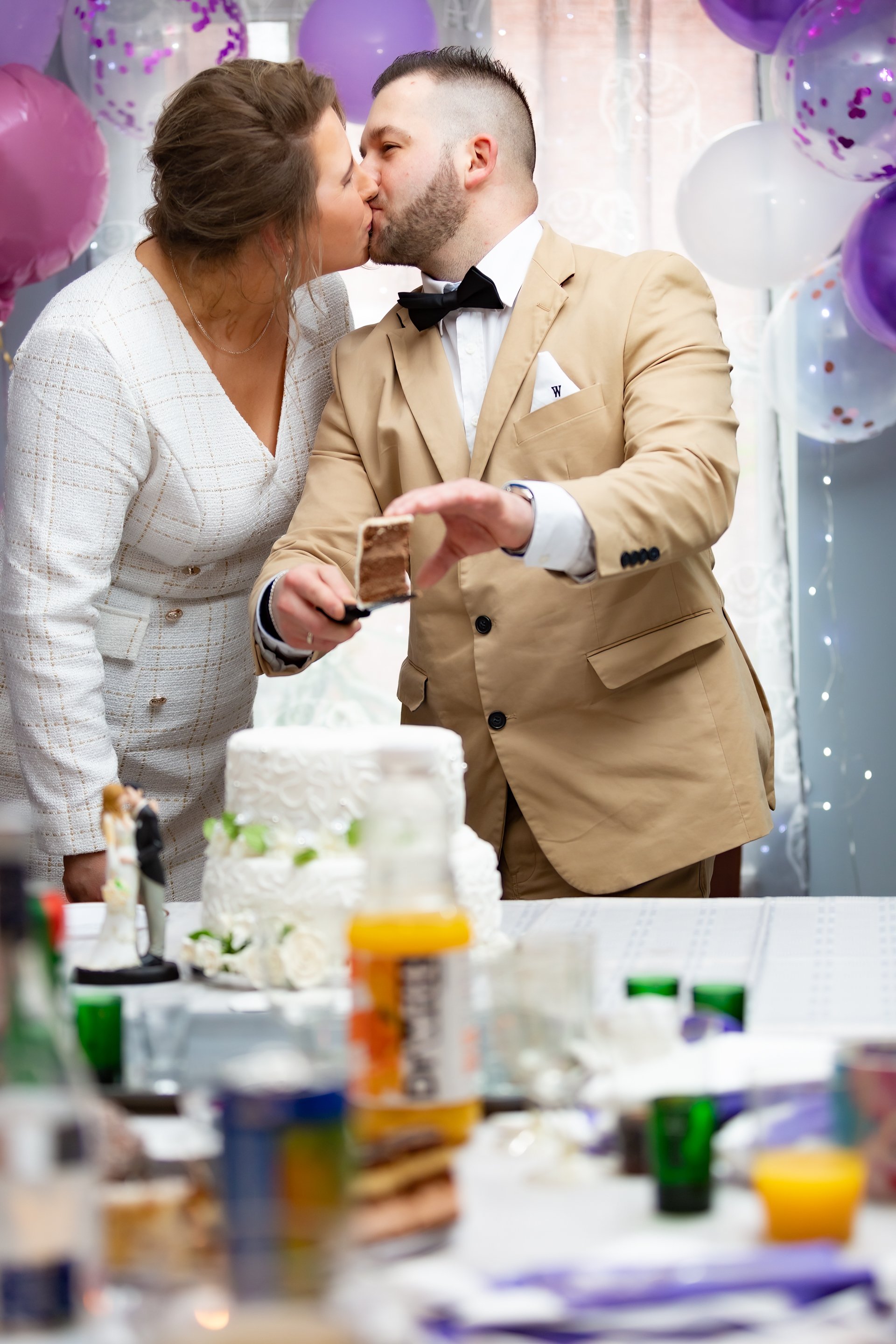 A newly wedded couple kisses while cutting a white, decorated cake at their wedding. Both are smartly dressed, surrounded by purple and white balloons, creating a joyful atmosphere.