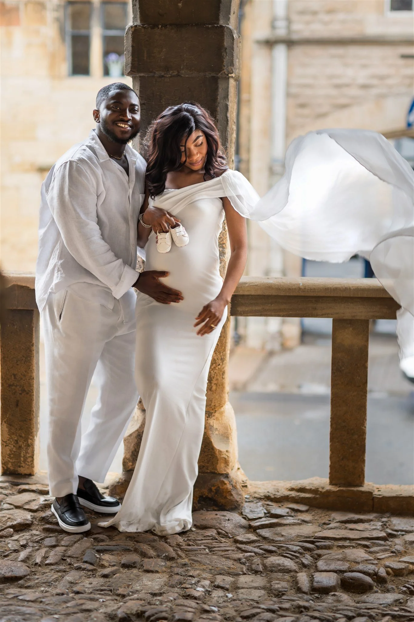 A couple in white attire poses lovingly on a stone balcony. The woman holds baby shoes on her baby bump, with her dress flowing elegantly in the breeze.