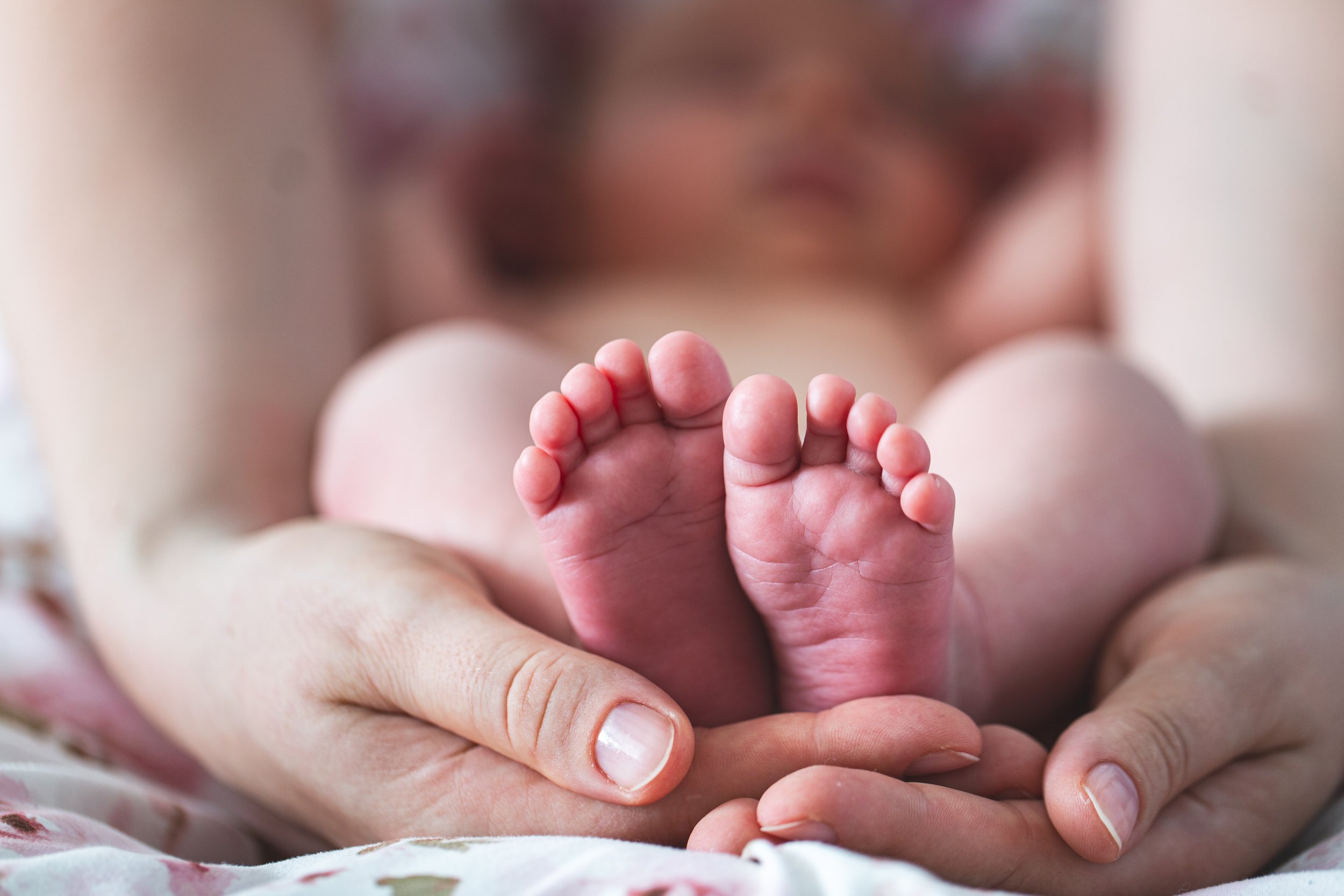 Close-up of a mother's hands gently cradling a baby's feet. The background is softly blurred, emphasizing a tender, nurturing, and loving moment.