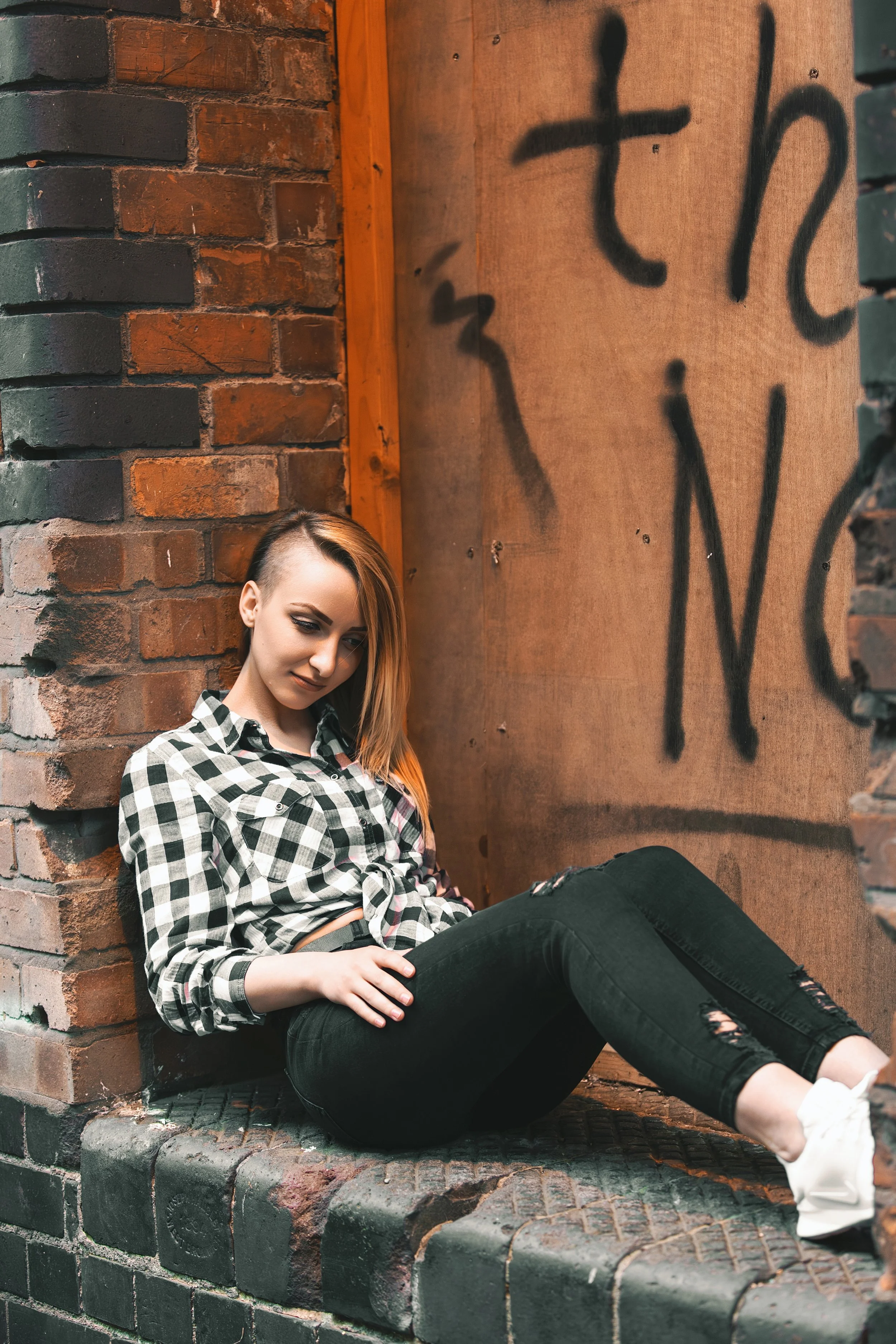 Portrait of a young woman with side-shaved hairstyle in black and white plaid shirt sits on a brick ledge, relaxed. Graffiti on wall. Urban, calm ambiance.