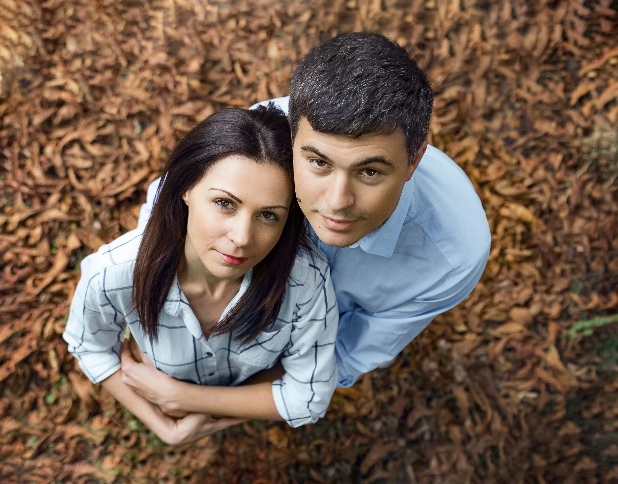Portrait of a couple stands closely on autumn leaves, looking up. The woman wears a striped shirt, while the man is in a blue shirt, evoking a serene, affectionate mood.