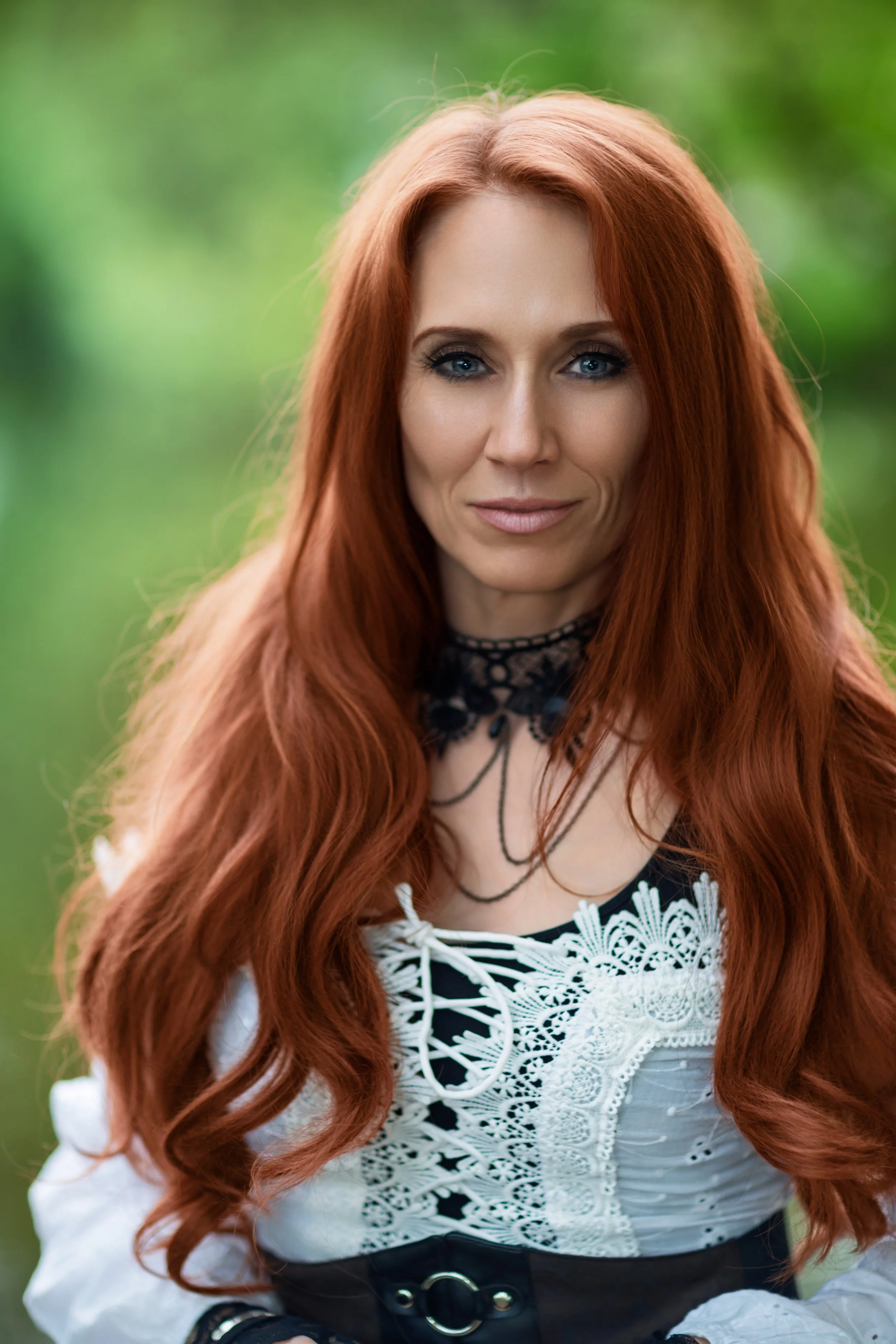 Portrait of a woman with long wavy red hair and a calm, confident expression wears a white lace corset and black choker, standing against a blurred green, natural background