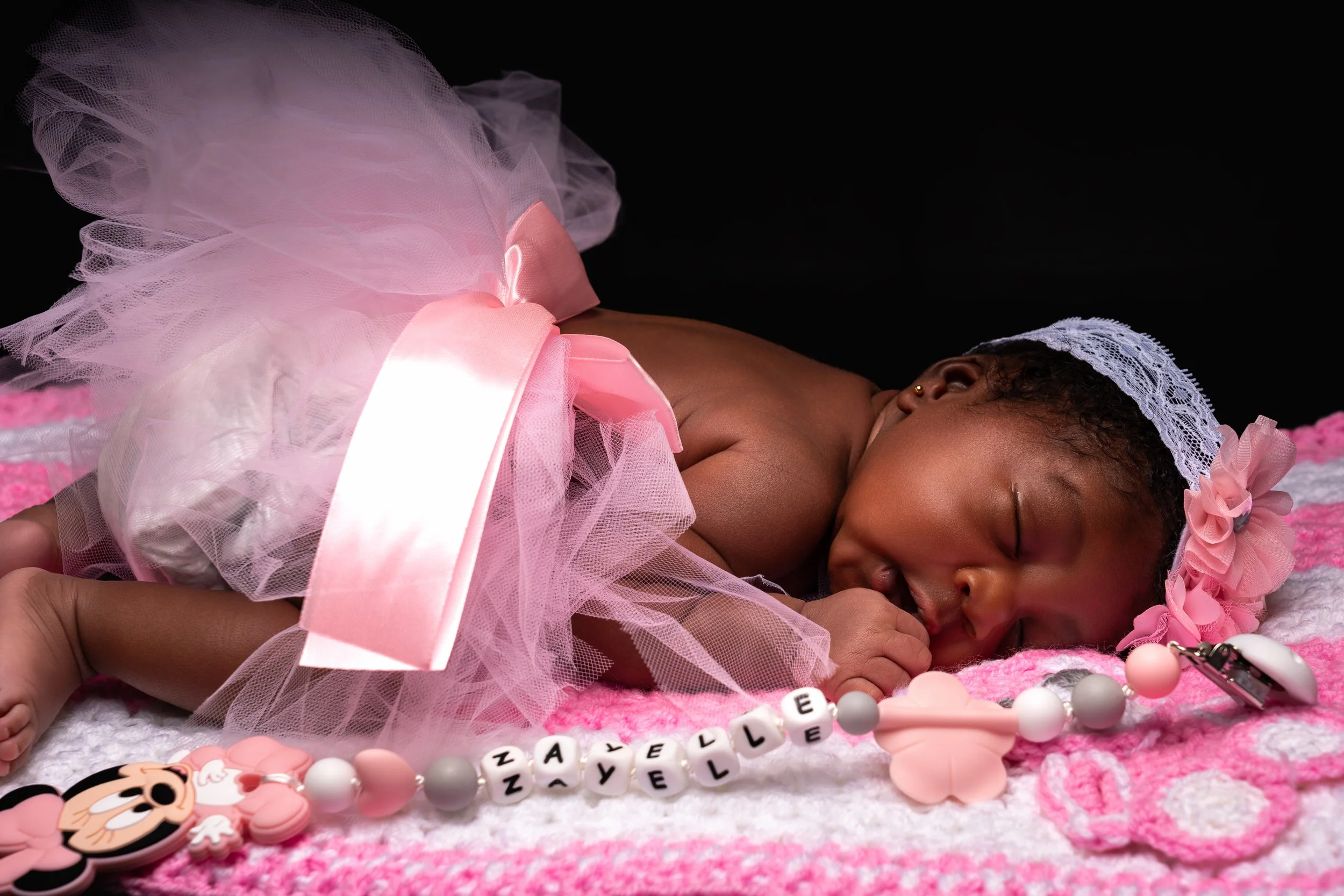 Sleeping baby in a pink tutu, resting on a pink and white crocheted blanket. A beaded name bracelet lies in front, creating a peaceful, tender scene.